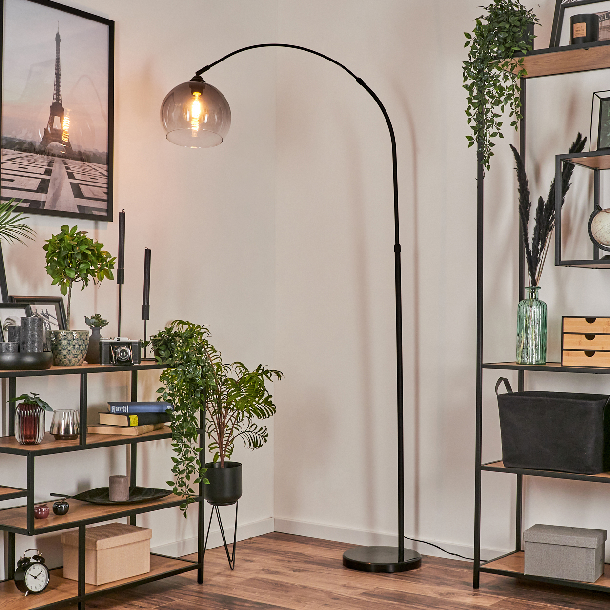 Eine Bogen-Stehlampe mit Glasschirm beleuchtet einen Raum. Regale zeigen Dekoration und Pflanzen. Ein Eiffelturm-Druck hängt an der Wand.