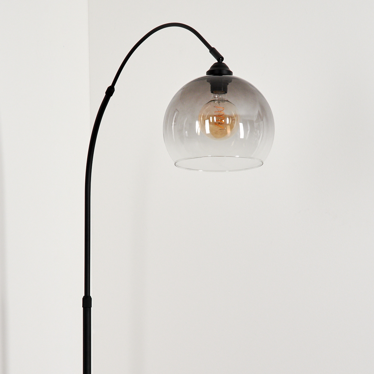 Eine schwarze Bogenstehlampe mit Glasschirm. Die Glühbirne ist sichtbar. Der Hintergrund ist eine weiße Wand. Die Lampe steht allein.