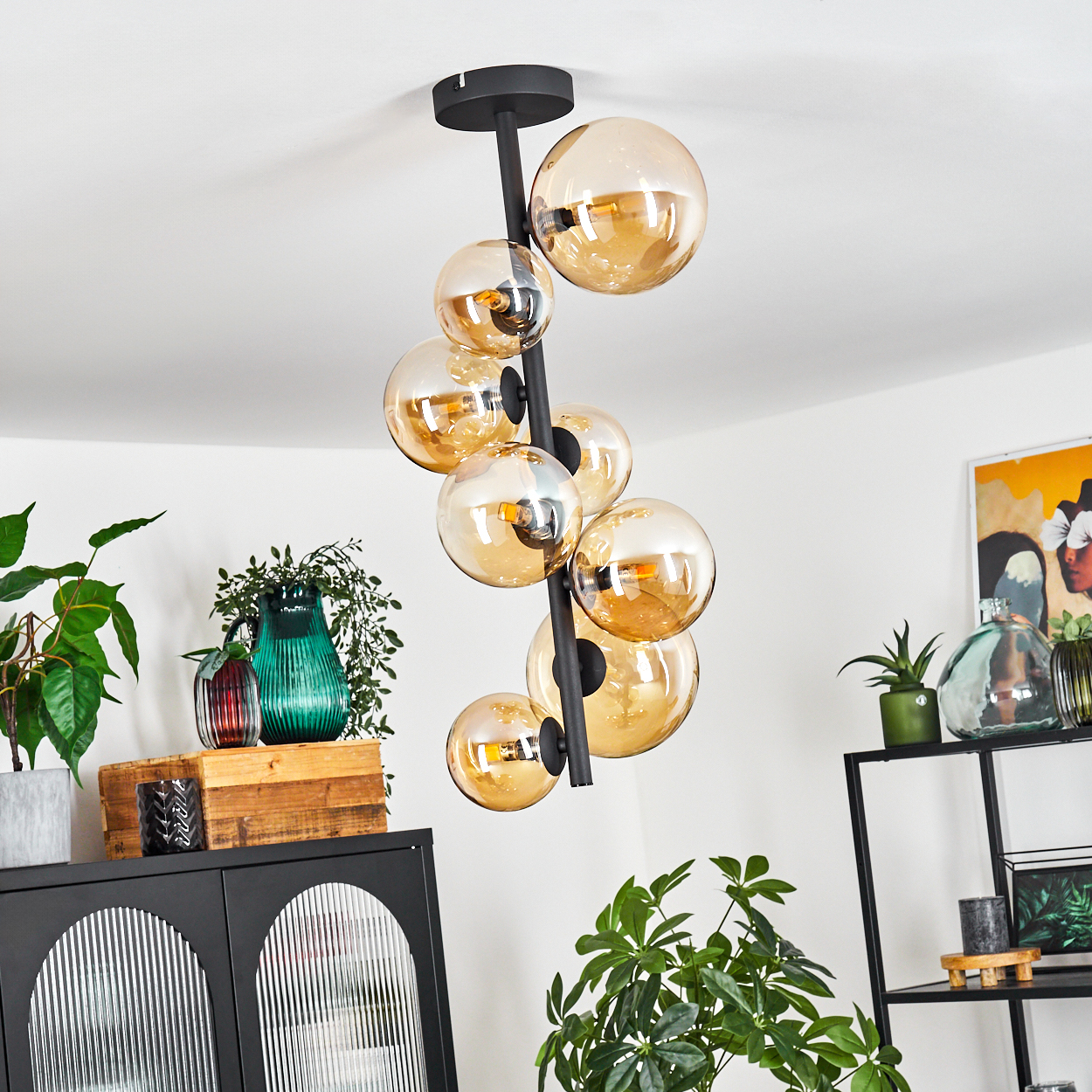 Eine schwarze Lampe mit vielen bernsteinfarbenen Kugeln hängt von der Decke. Darunter ein Holzschrank mit Pflanzen und Vasen.