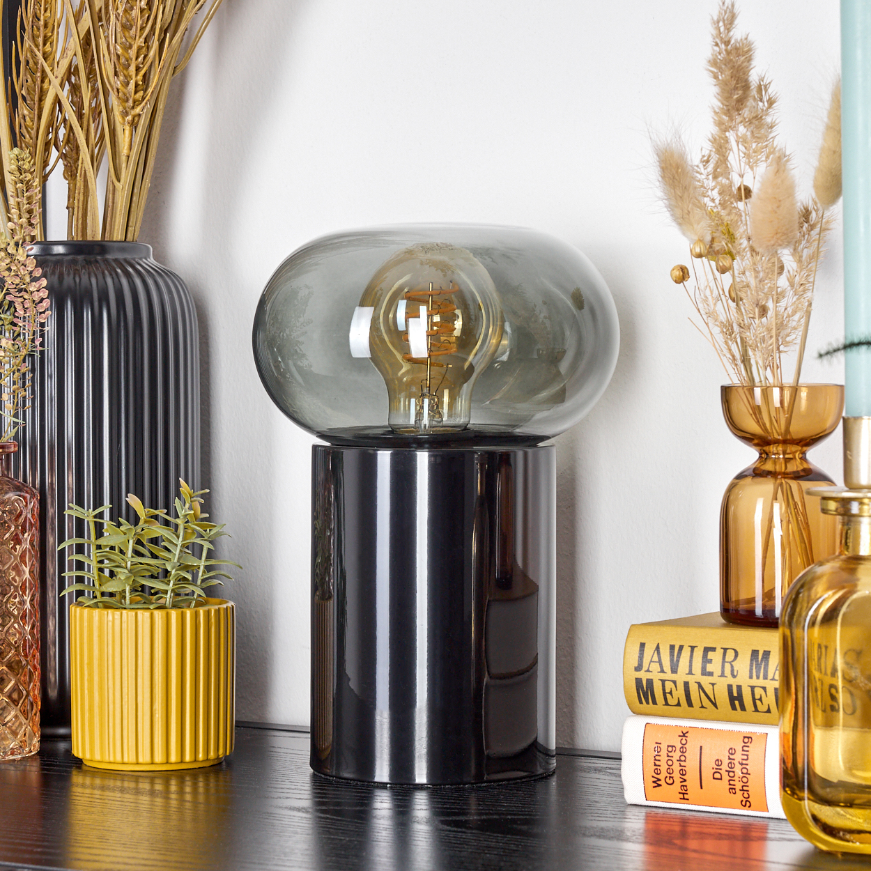 Eine moderne Lampe mit grauem Glasschirm und schwarzem Sockel steht auf einem Regal mit Pflanzen, Vasen und Büchern. Warme Farben dominieren.