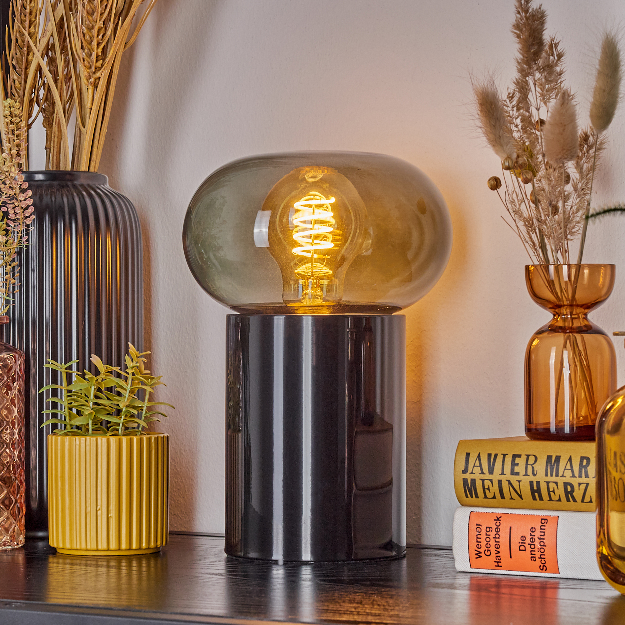 Eine beleuchtete Lampe mit schwarzem Sockel und transparentem Glasschirm. Verschiedene Vasen mit Trockenblumen und Bücher sind darum platziert.