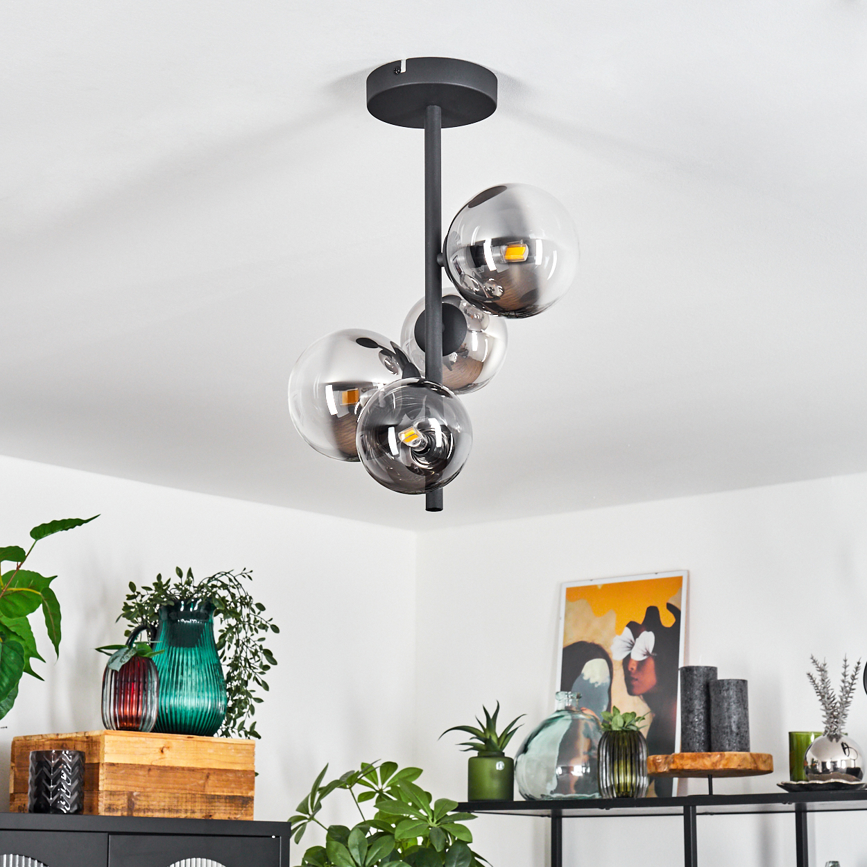 Eine moderne Hängelampe mit vier Glaskugeln hängt von der Decke. Vasen, Pflanzen und Kunstwerke schmücken ein Regal darunter. Die Szene ist hell.