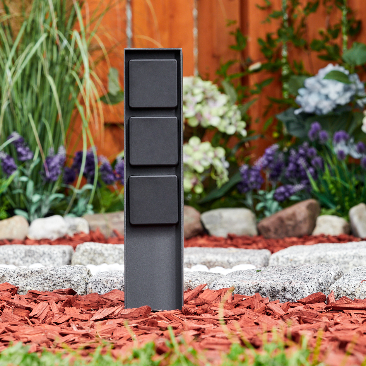 Ein schwarzer Metallpfosten mit drei quadratischen Abschnitten steht in einem Gartenbeet mit roter Mulch, Steinen und Pflanzen im Hintergrund.