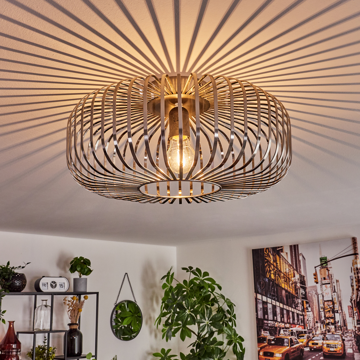 Eine moderne Deckenlampe mit käfigartigem Schirm. Sie wirft Schattenmuster an die Decke. Pflanzen und ein Stadtbild-Gemälde sind im Raum.