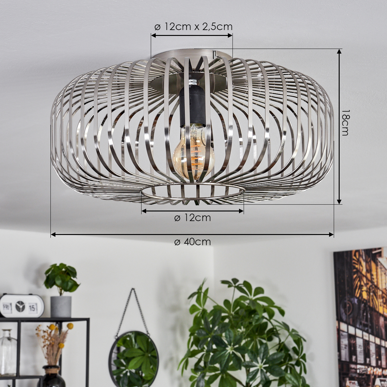 Eine silberne, käfigartige Deckenlampe mit sichtbarer Glühbirne. Das Bild zeigt auch Pflanzen, eine Uhr, einen Spiegel und ein Gemälde.