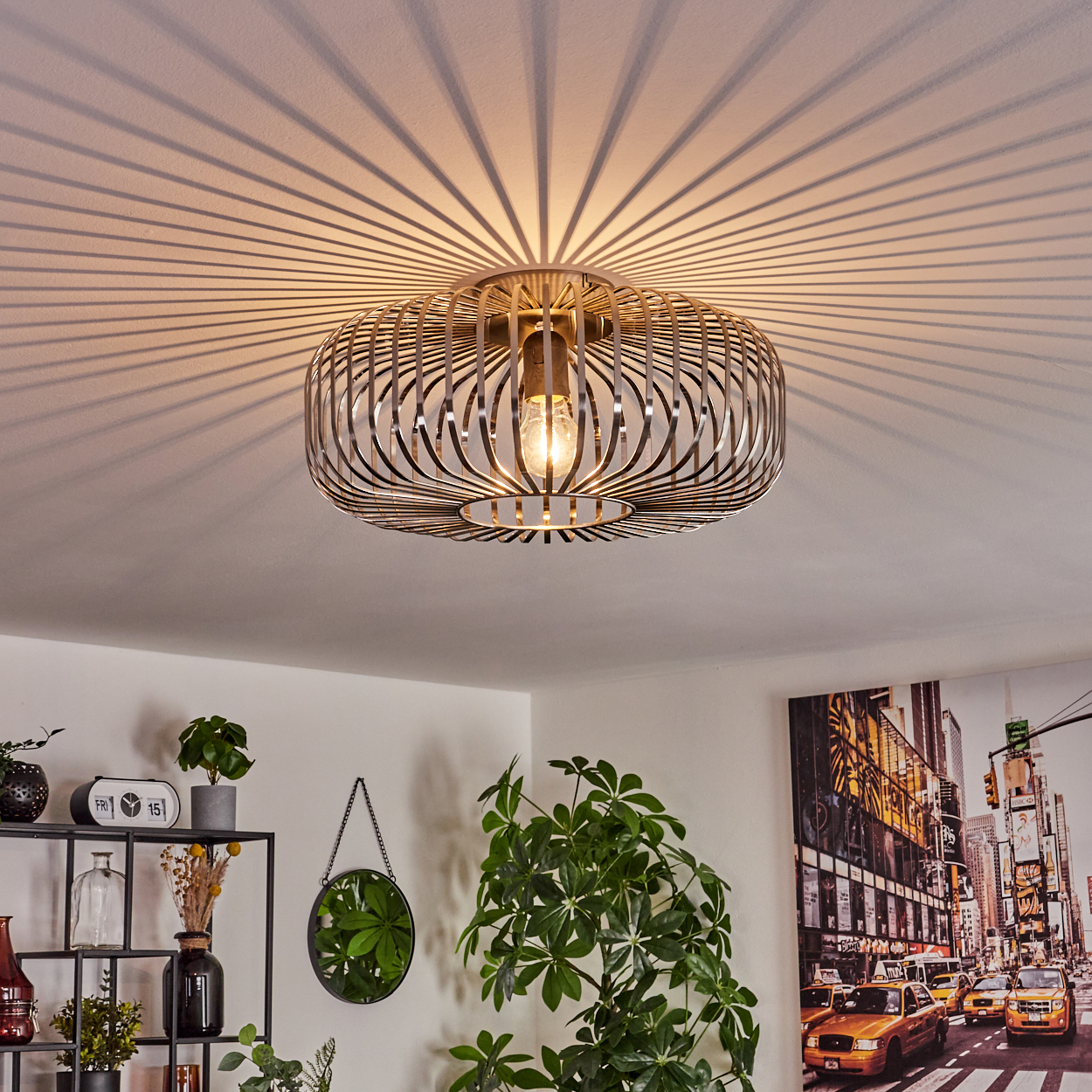 Eine moderne Deckenlampe wirft radiale Schatten. Darunter ein Regal mit Deko, ein Spiegel und ein New York City-Kunstwerk. Grüne Pflanzen.