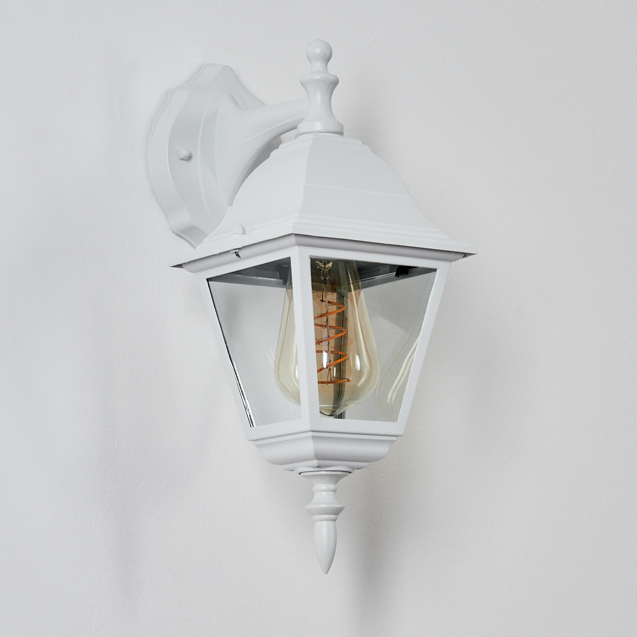 Eine weiße Wandlaterne. Sie hat eine klare Glasumhüllung mit einer Glühbirne im Vintage-Stil. Der Hintergrund ist eine schlichte weiße Wand.