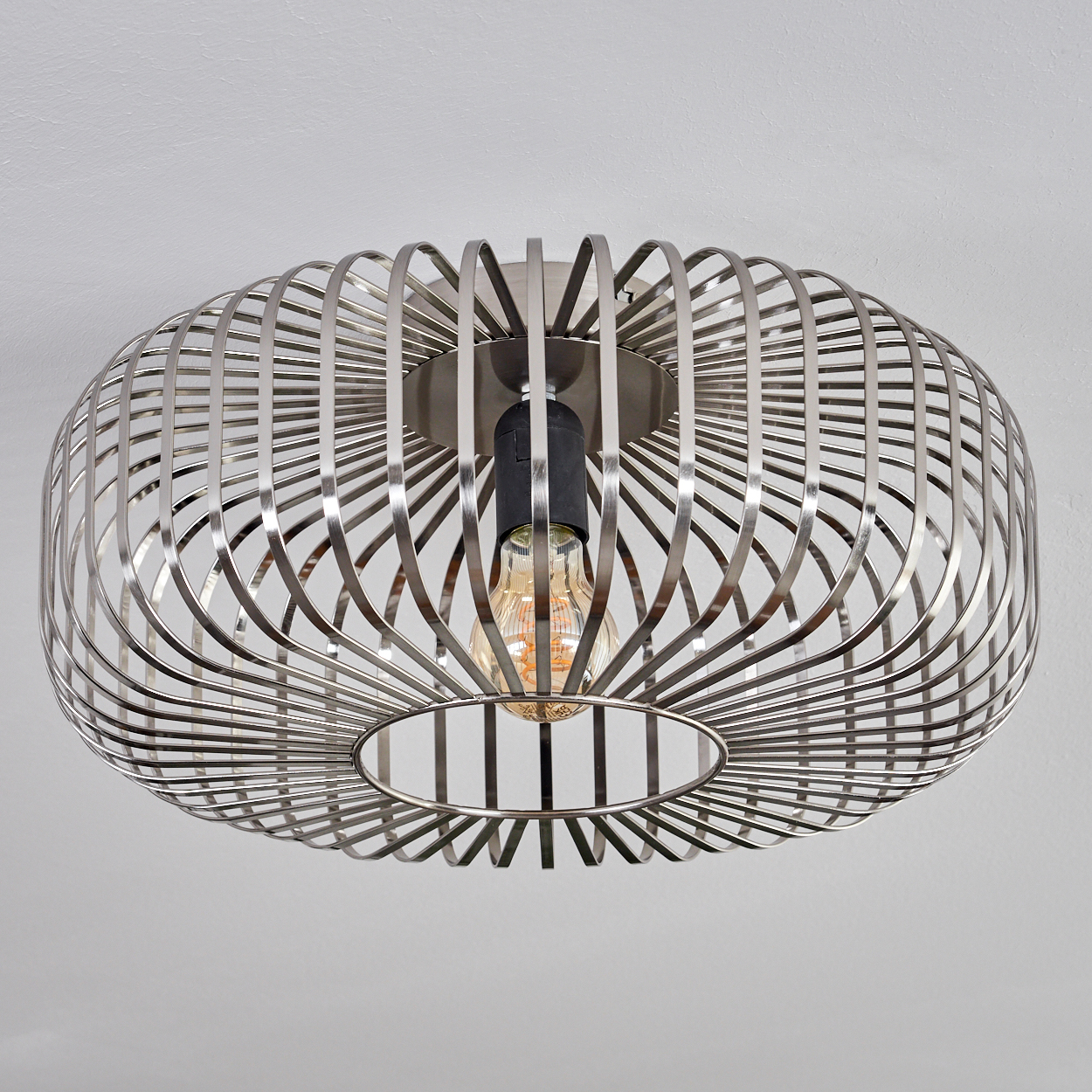 Eine silberne Metallkäfig-Leuchte mit sichtbarer Glühbirne ist an einer weißen Decke montiert. Der Käfig hat eine ovale Form.