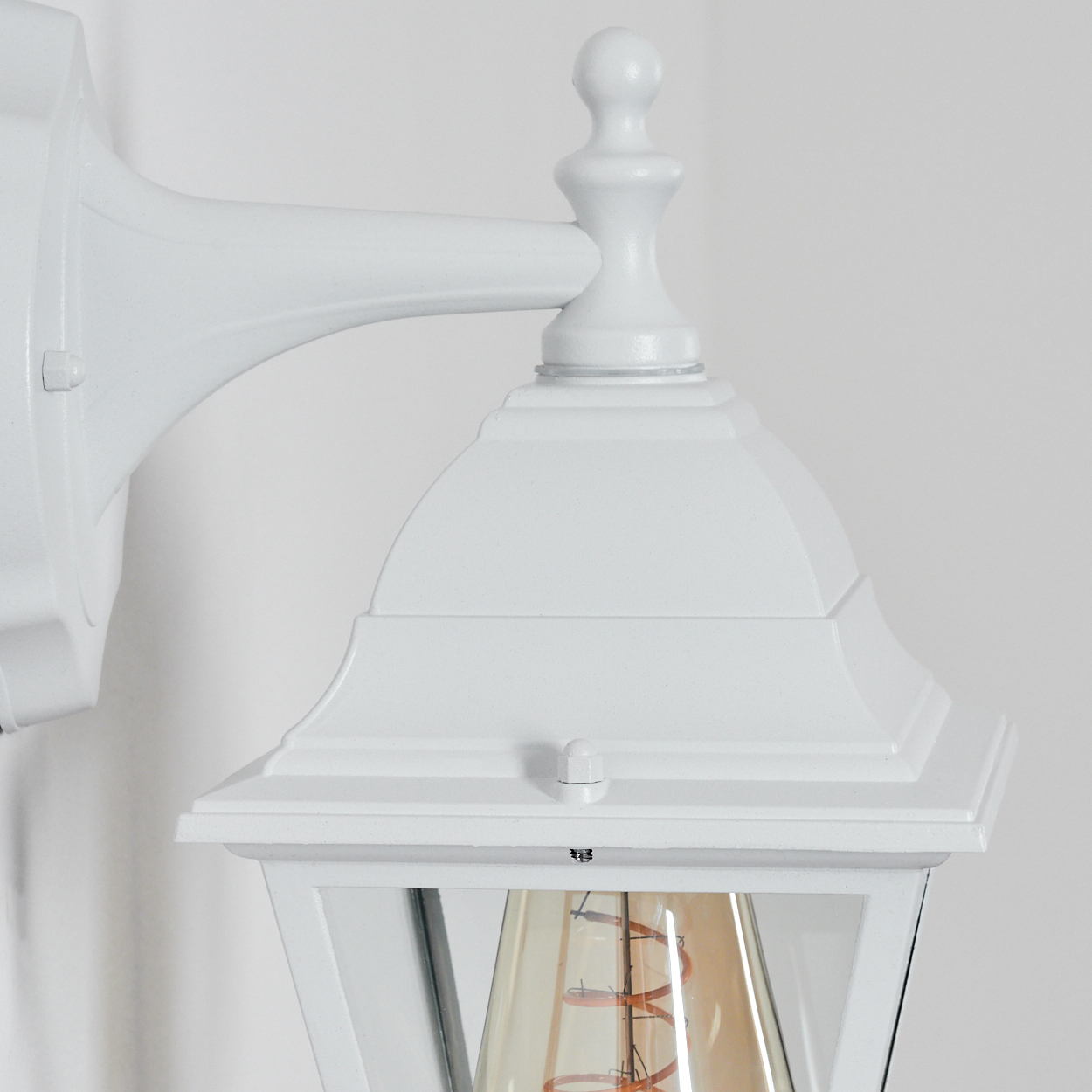 Nahaufnahme einer weißen Außenlampe. Die Lampe hat ein Glasgehäuse und eine sichtbare Glühbirne. Der Hintergrund ist eine weiße Wand.