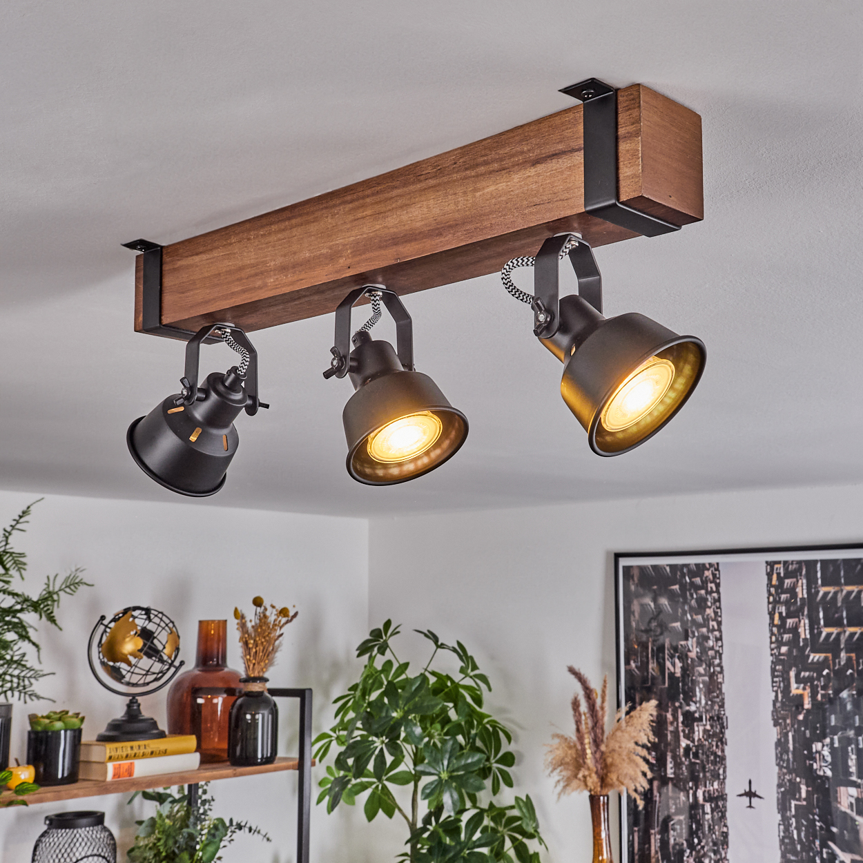 Drei schwarze Strahler an einem Holzbalken an einer weißen Decke. Darunter ein Regal mit Dekor und Pflanzen.