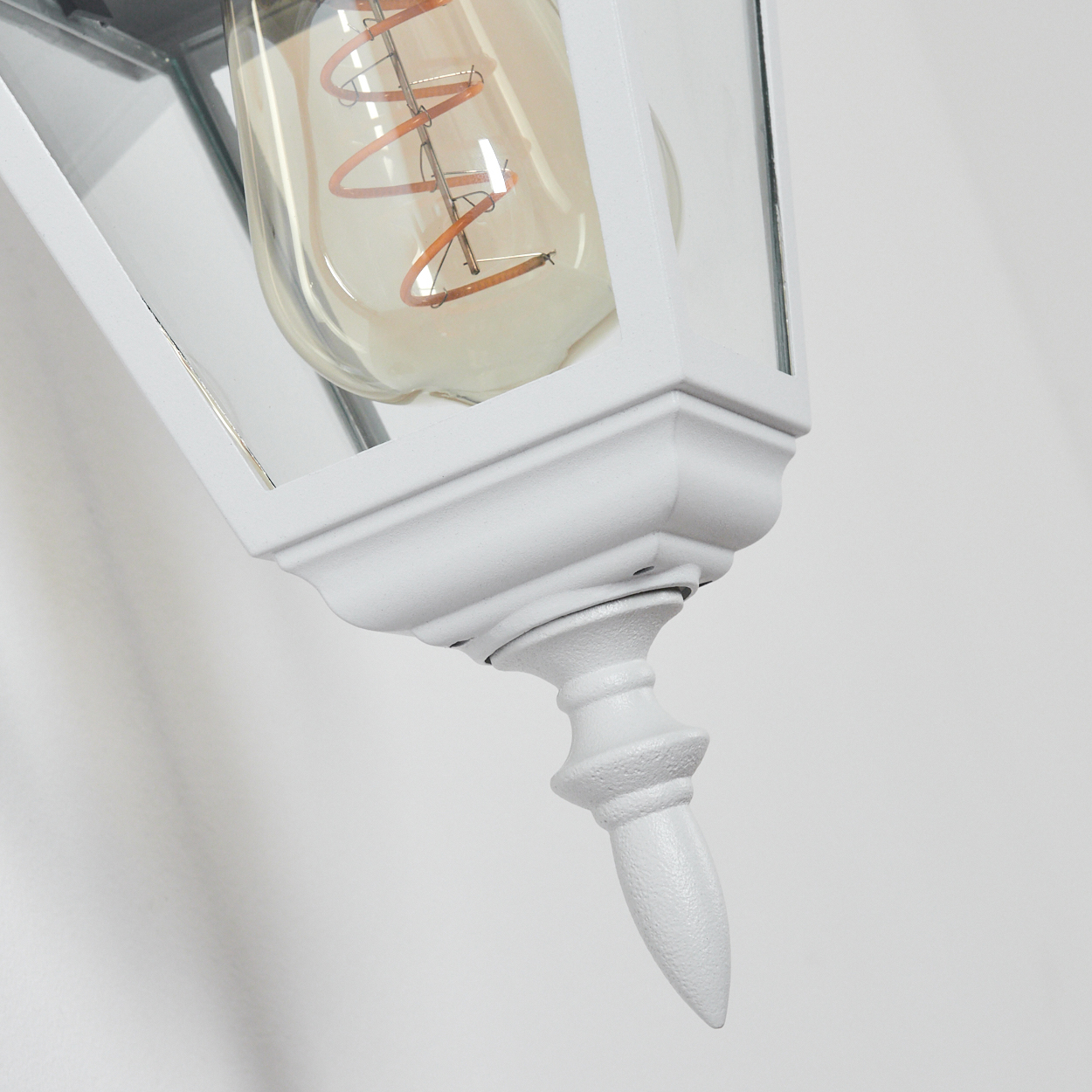 Nahaufnahme einer weißen, verzierten Außenlampe mit sichtbarer Glühbirne. Der Hintergrund ist eine schlichte, weiße Wand.
