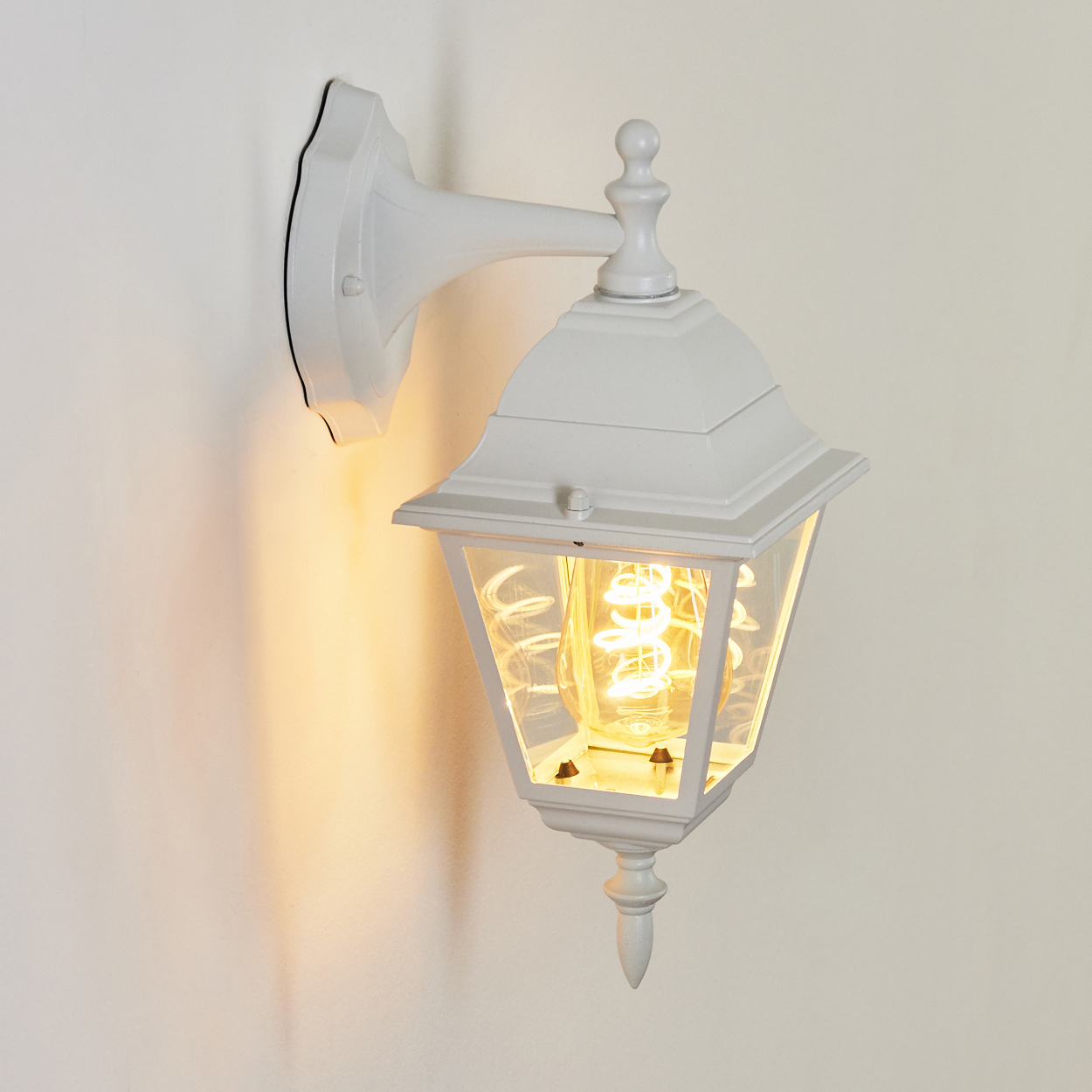 Eine weiße Außenlampe ist an einer Wand befestigt. Sie hat ein Glasgehäuse und eine helle, spiralförmige Glühbirne.