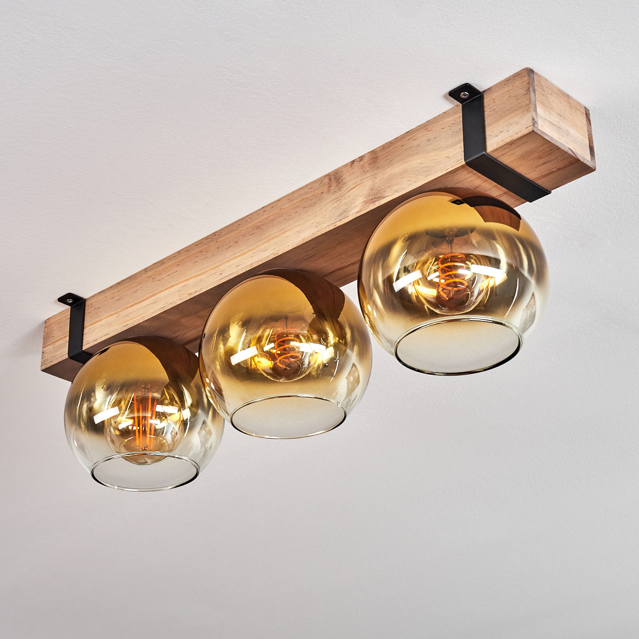 Ein Holzbalken mit drei goldenen Glaskugelleuchten. Jede Leuchte hat einen sichtbaren Glühfaden. Schwarze Metallhalterungen halten den Balken.