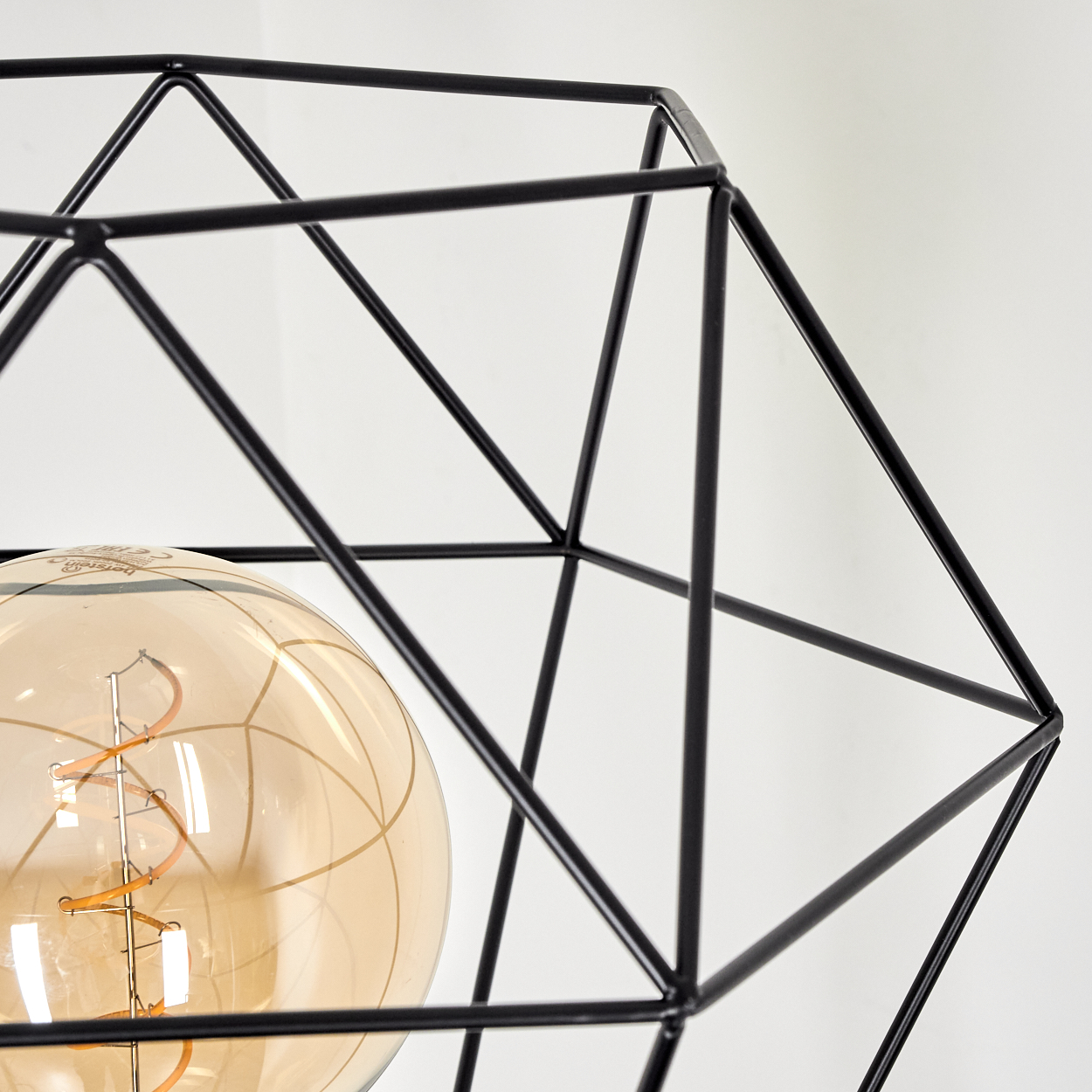 Nahaufnahme eines schwarzen geometrischen Lampenschirms. Im Inneren befindet sich eine runde, bernsteinfarbene Glühbirne. Der Hintergrund ist eine weiße Wand.