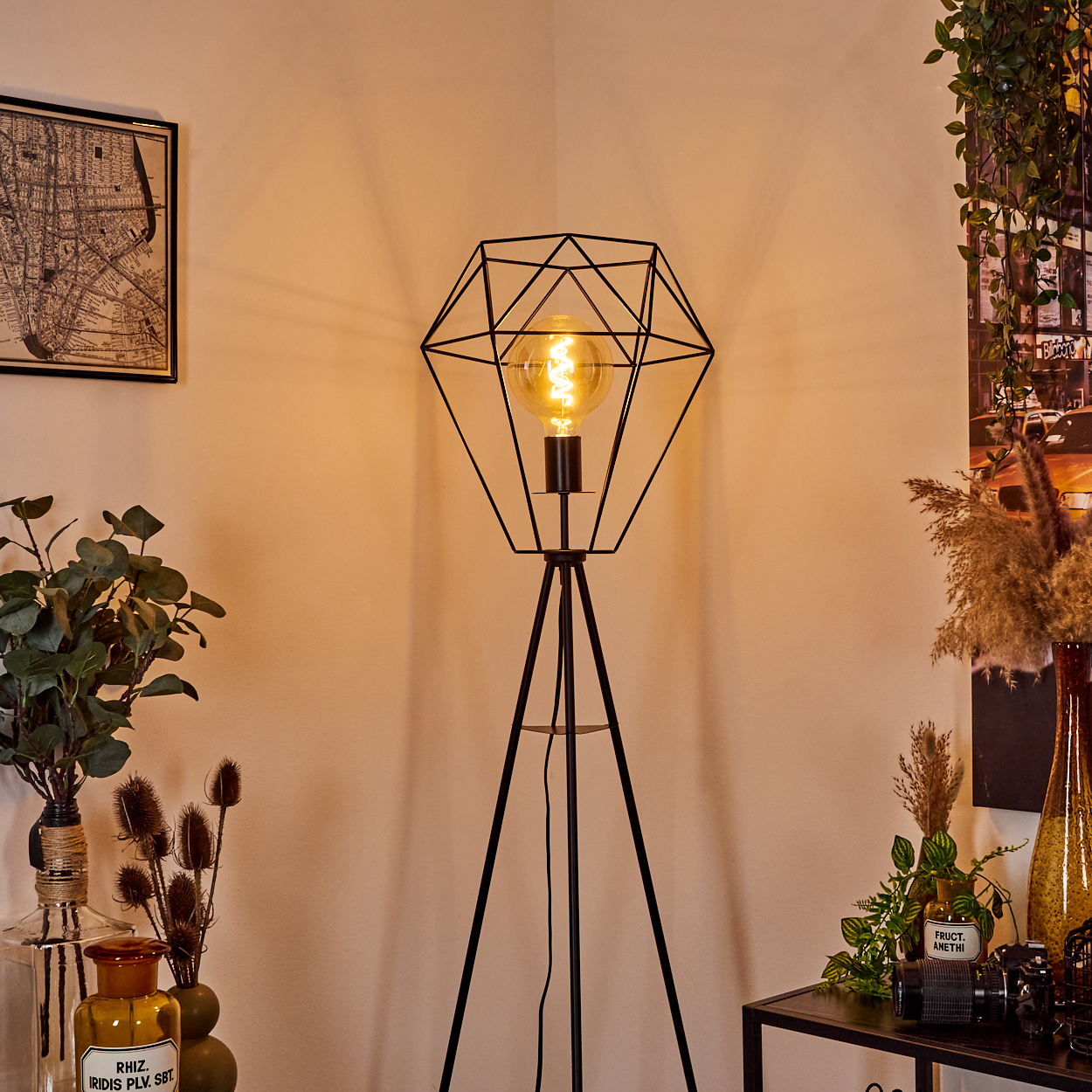 Eine schwarze, geometrische Stehlampe mit einer leuchtenden Glühbirne steht in einer Ecke. Pflanzen und ein Kartenbilderrahmen sind ebenfalls vorhanden.