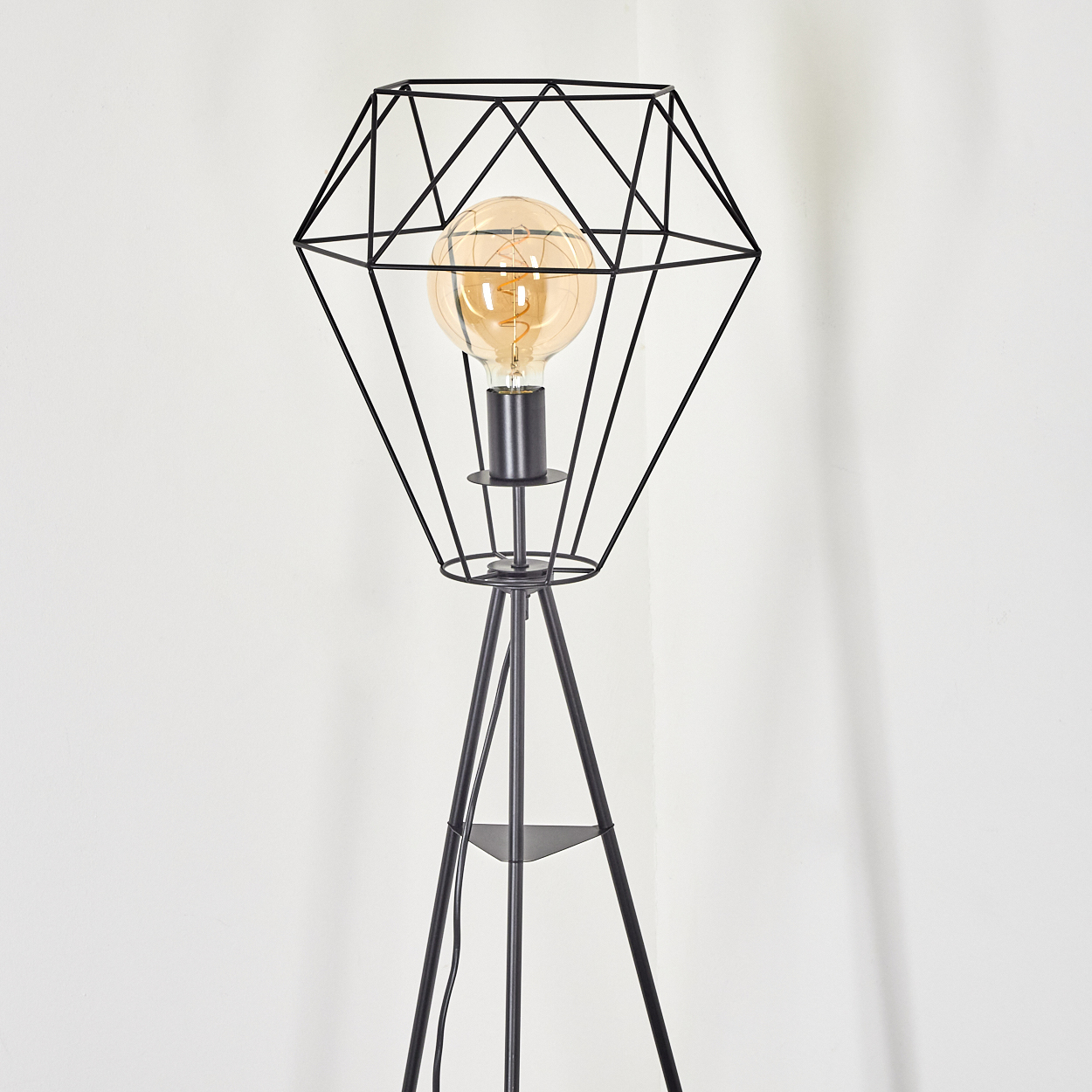 Eine schwarze geometrische Stehlampe mit rautenförmigem Schirm und warm getönter Glühbirne vor weißer Wand.