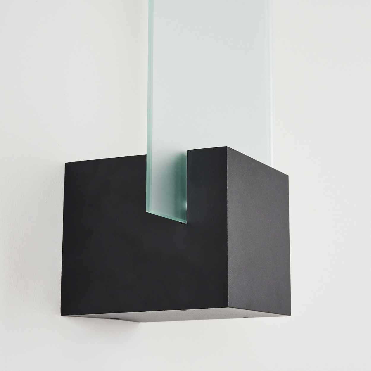 Eine schwarze, quadratische Wandleuchte mit einer nach oben ragenden Milchglasscheibe an einer weißen Wand.