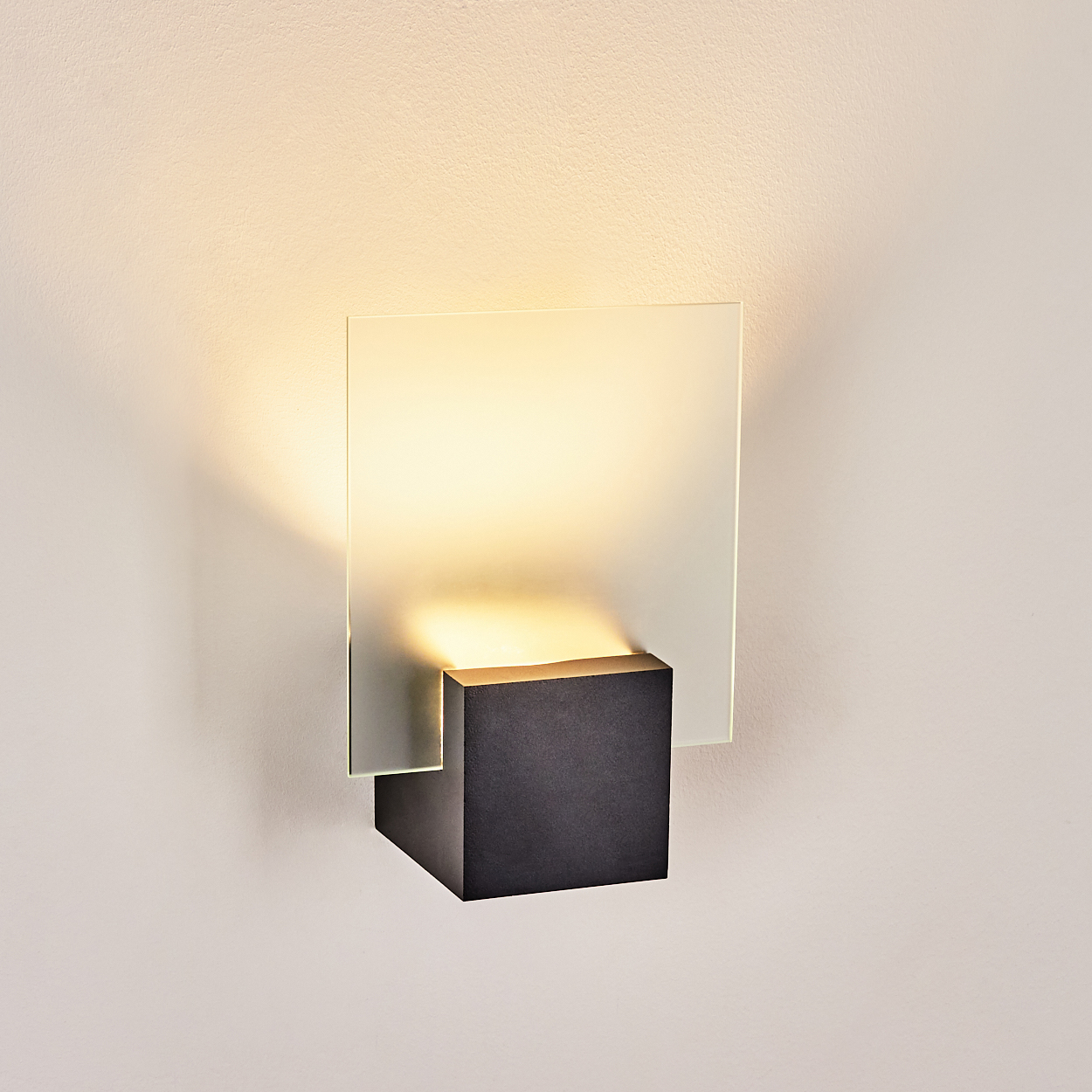 Eine moderne Wandleuchte mit quadratischem Milchglas und schwarzem quadratischem Sockel. Sie ist an einer weißen Wand montiert und strahlt warmes Licht aus.
