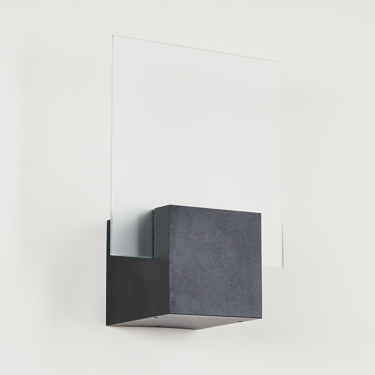 Moderne Wandleuchte mit quadratischem, dunkelgrauem Block, klarer Glasscheibe und schwarzer Montageplatte an weißer Wand.