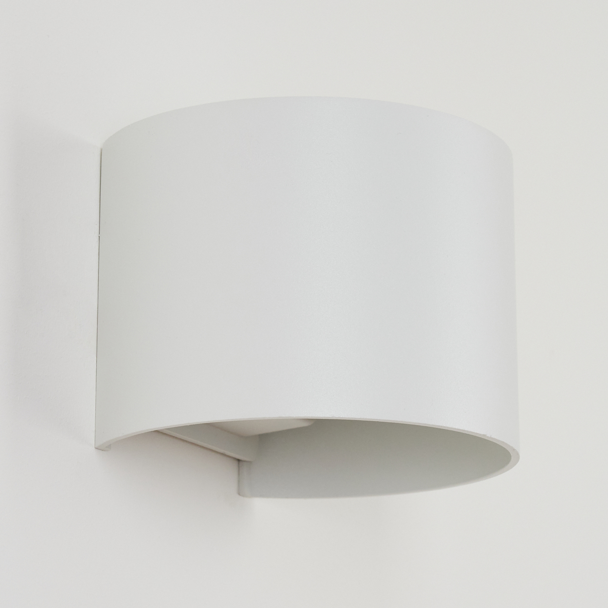 Eine weiße, halb zylindrische Wandleuchte ist an einer weißen Wand montiert. Die Lampe hat eine solide, gebogene Form mit offenem Boden.
