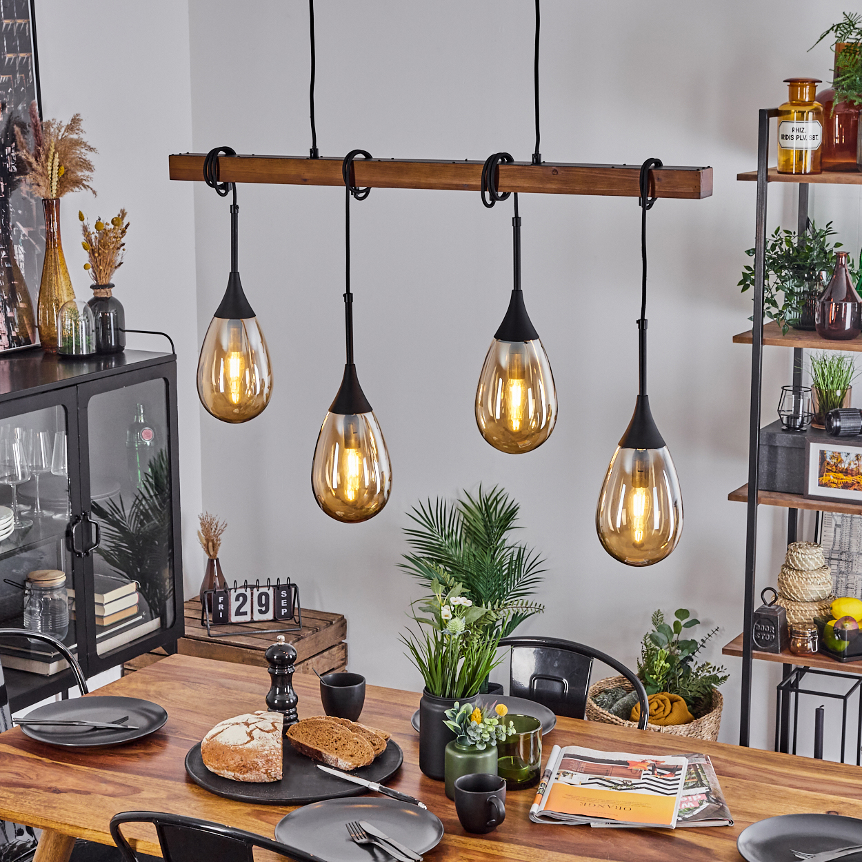 Esszimmer mit Holztisch, schwarzen Stühlen und Pendelleuchten. Brot und Teller auf dem Tisch, Regale im Hintergrund.