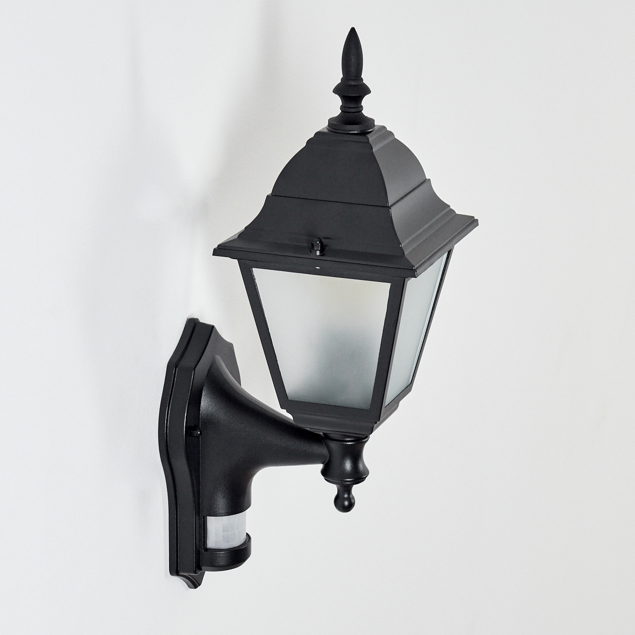 Eine schwarze, an der Wand montierte Außenlampe mit einer Milchglasscheibe. Sie hat einen Bewegungssensor und ist an einer weißen Wand befestigt.