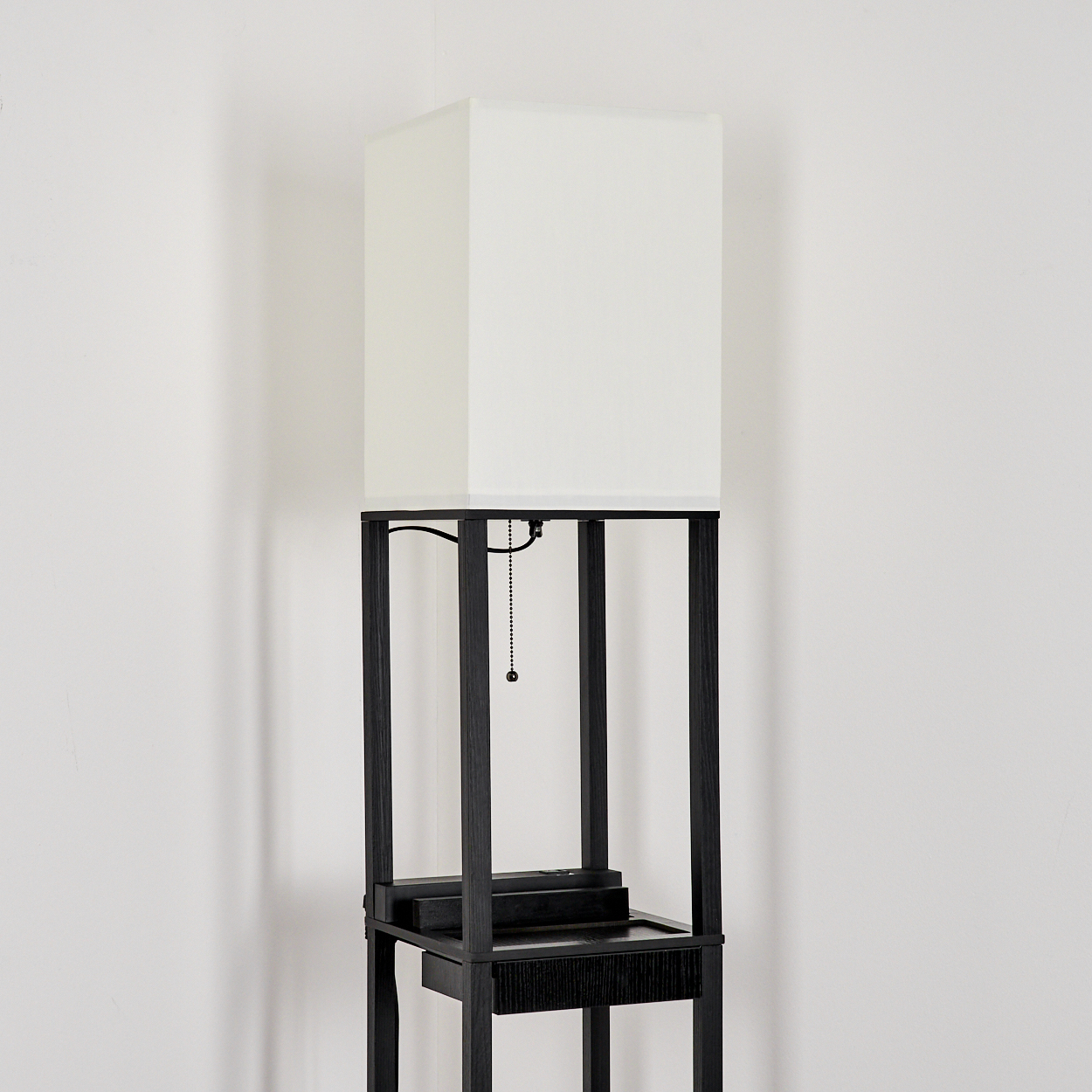 Eine hohe, schwarz gerahmte Lampe mit einem weißen, rechteckigen Schirm. Die Lampe hat eine kleine Schublade und steht vor einer weißen Wand.