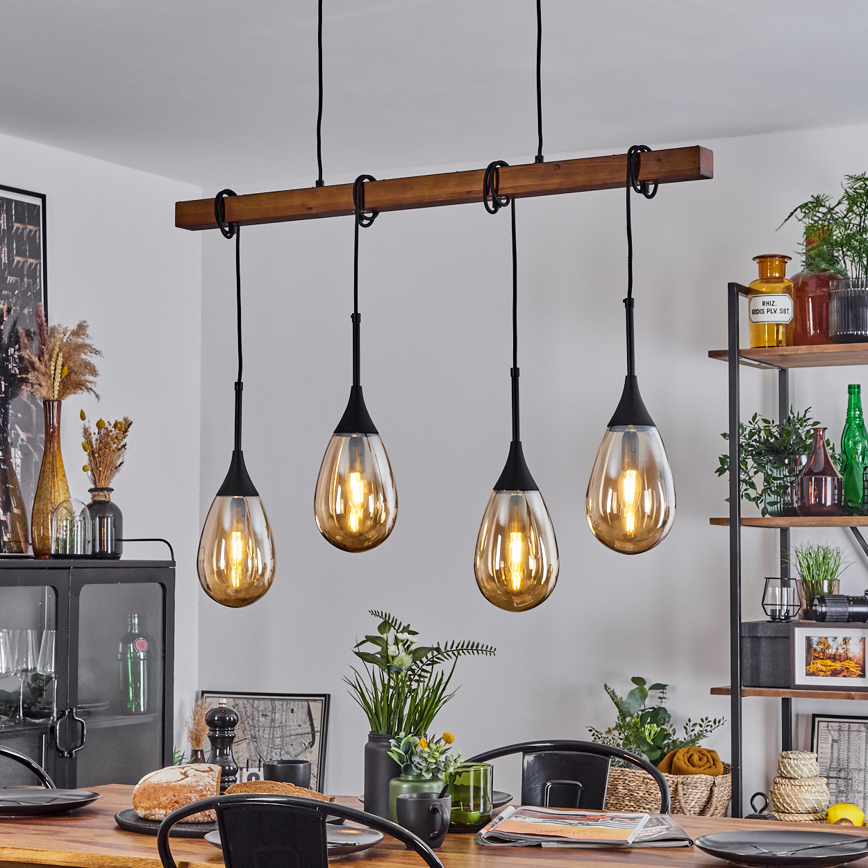 Vier Pendelleuchten mit bernsteinfarbenen Glasschirmen hängen über einem Holztisch, der für eine Mahlzeit gedeckt ist. Regale mit Pflanzen sind im Hintergrund.