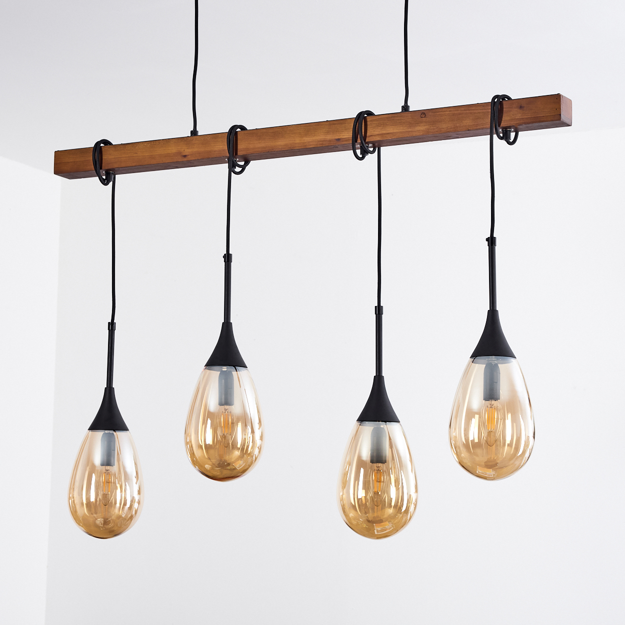 Vier Pendelleuchten mit bernsteinfarbenen, tropfenförmigen Glasschirmen hängen an einem Holzbalken. Schwarze Kabel und Armaturen vor weißer Wand.