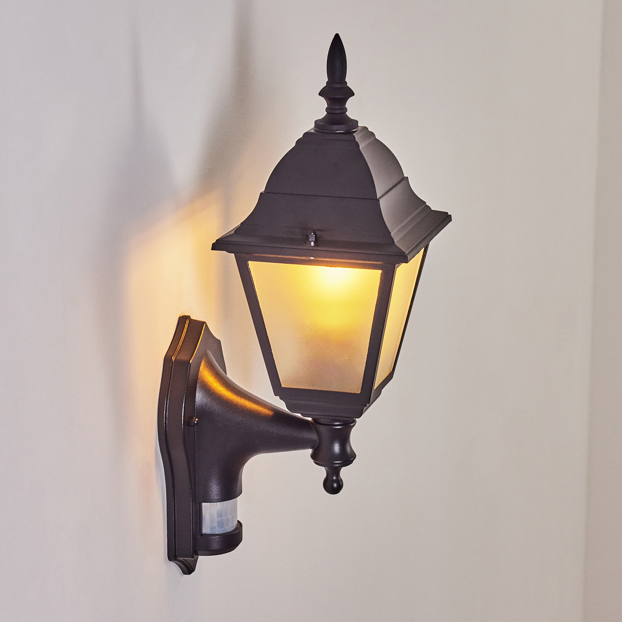 Eine schwarze Außenlampe mit Milchglas, wandmontiert. Das Licht ist an und wirft einen warmen Schein, mit einem Bewegungssensor darunter.