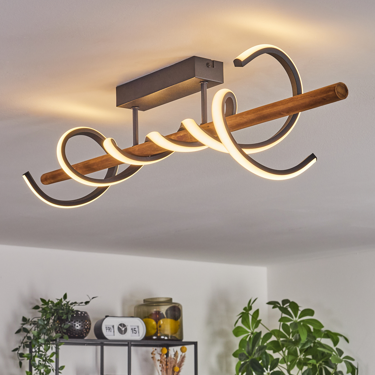 Eine moderne Deckenleuchte mit Holzbalken und wirbelnden, beleuchteten schwarzen Metallakzenten. Pflanzen und Deko im Hintergrund.