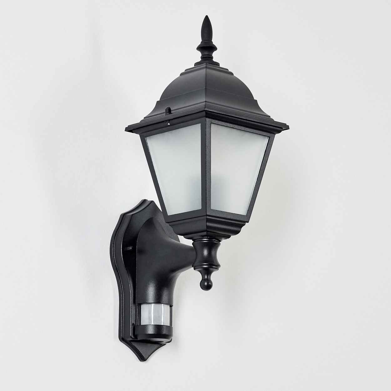 Eine schwarze Außenwandleuchte mit Bewegungssensor ist an einer weißen Wand montiert. Die Lampe hat ein klassisches Design.