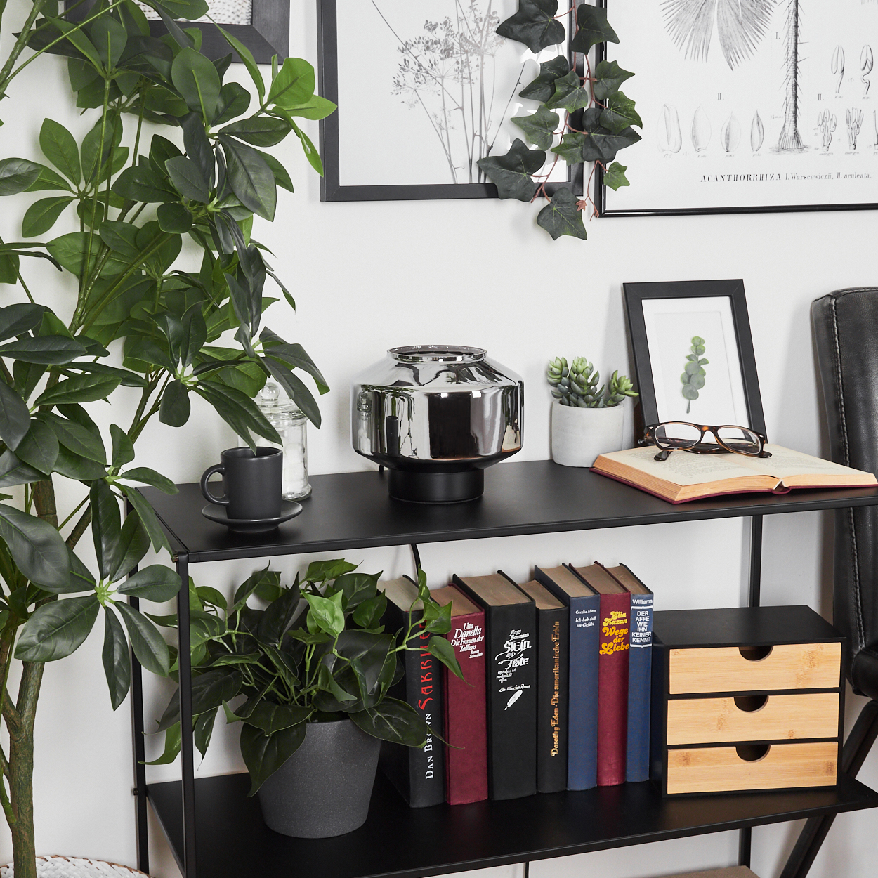 Ein schwarzes Regal mit Büchern, Pflanzen und einer Lampe. Bilder hängen an der weißen Wand. Auf dem Regal befinden sich eine Tasse und eine Brille.