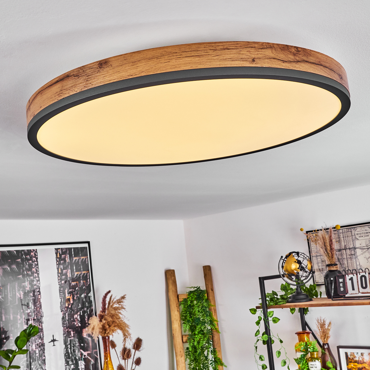 Ovale Deckenleuchte mit Holzrahmen und schwarzem Rand. Darunter ein Wohnzimmer mit Dekoration: Leiter, Pflanzen, Bilder.