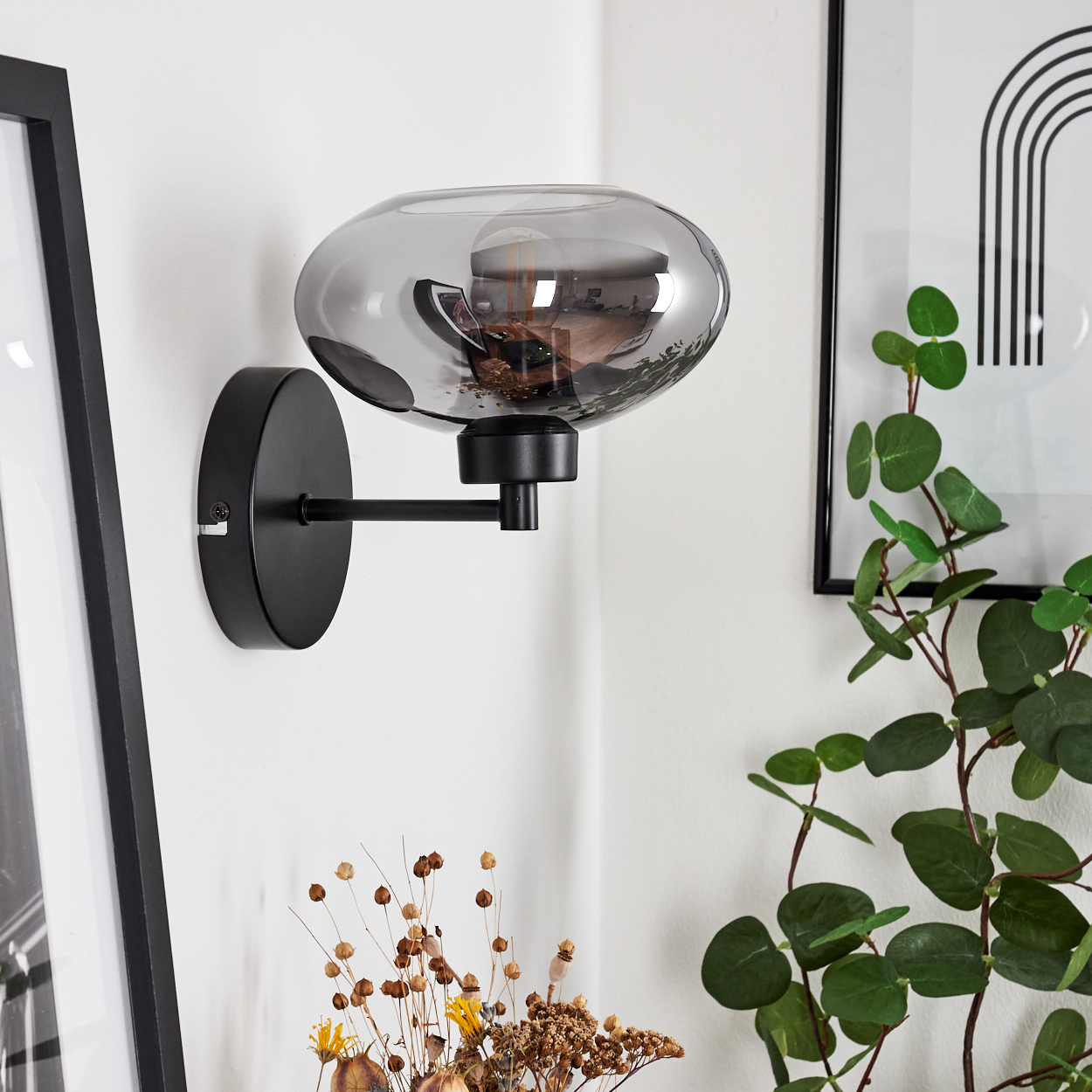 Eine schwarze Wandlampe mit grauem Glasschirm ist an einer weißen Wand montiert. Ein gerahmtes Bild und Pflanzen sind ebenfalls zu sehen.