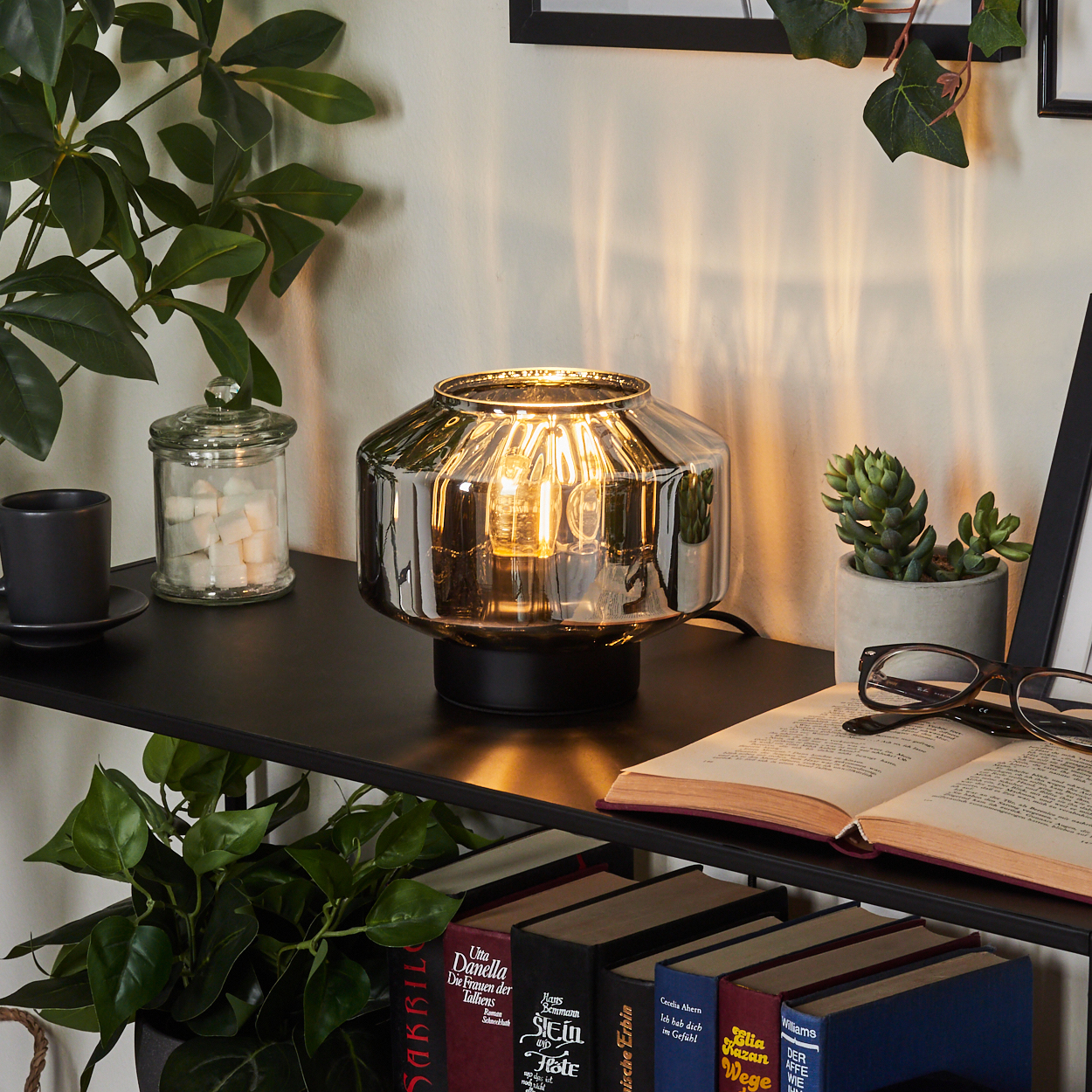 Eine beleuchtete Lampe steht auf einem schwarzen Regal mit Büchern, einer Pflanze und einer Tasse. Ein Glas mit Zuckerwürfeln und Gläser sind ebenfalls vorhanden.