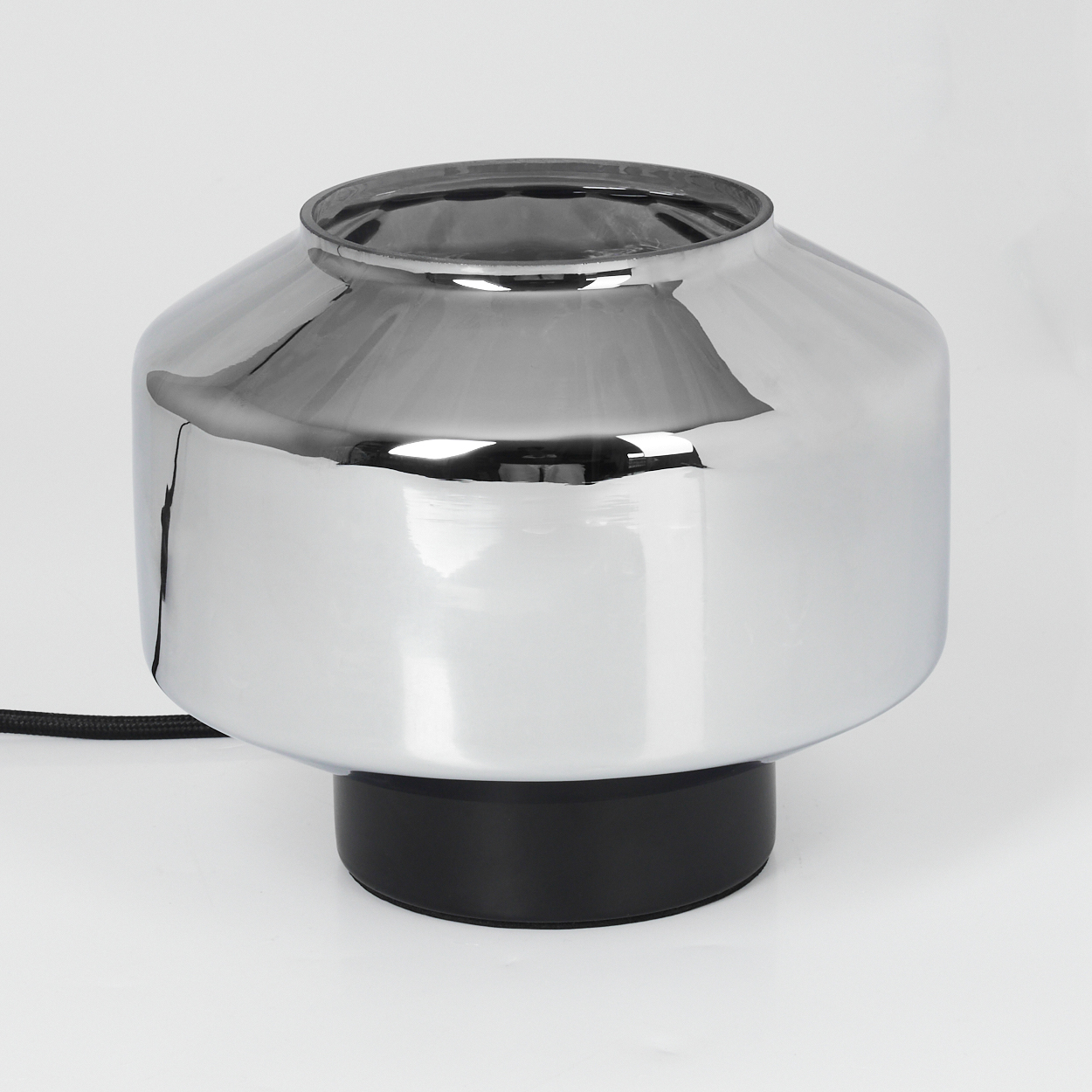 Eine Chromlampe mit schwarzem Fuß steht auf einer weißen Oberfläche. Die Lampe ist reflektierend, ein schwarzes Netzkabel ist sichtbar.