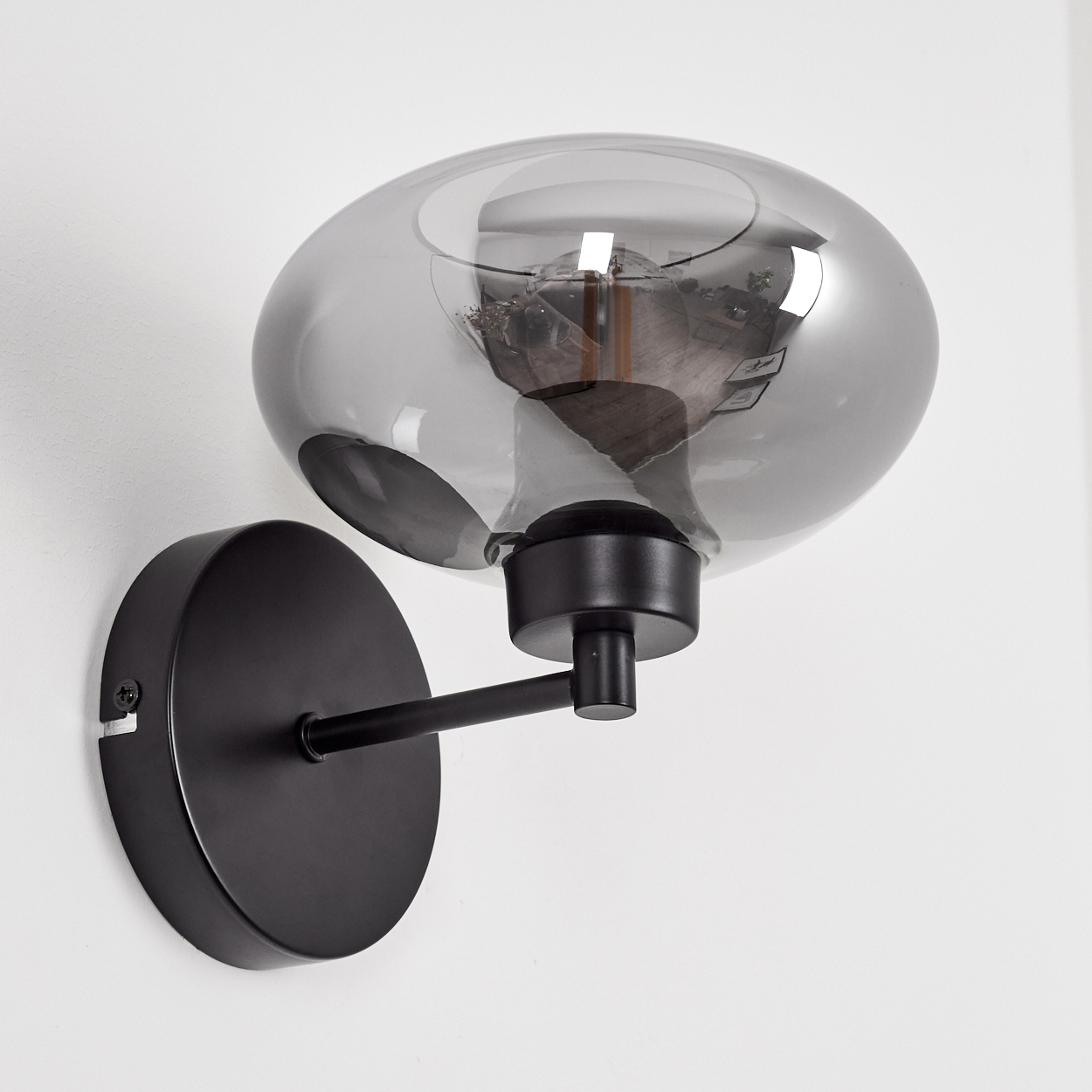Eine schwarze Wandleuchte mit rauchfarbenem Glasschirm. Die Lampe ist an einer weißen Wand befestigt. Die Glühbirne ist im Schirm sichtbar.