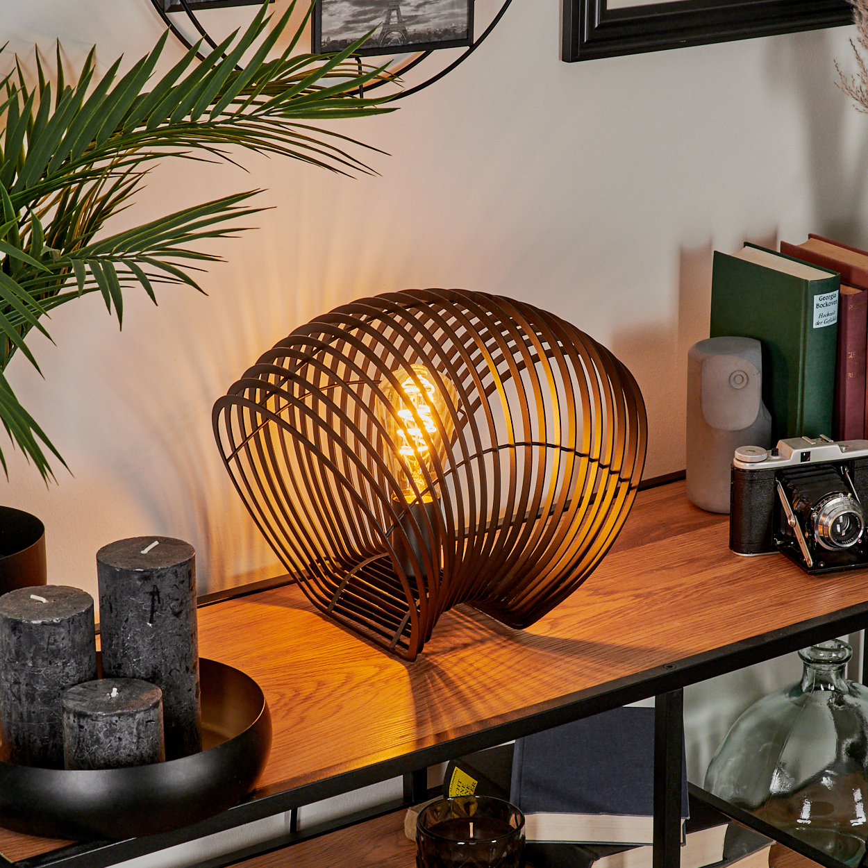Eine dekorative Tischlampe mit warmem Licht, Kerzen, Büchern und einer Kamera auf einem Holzregal.