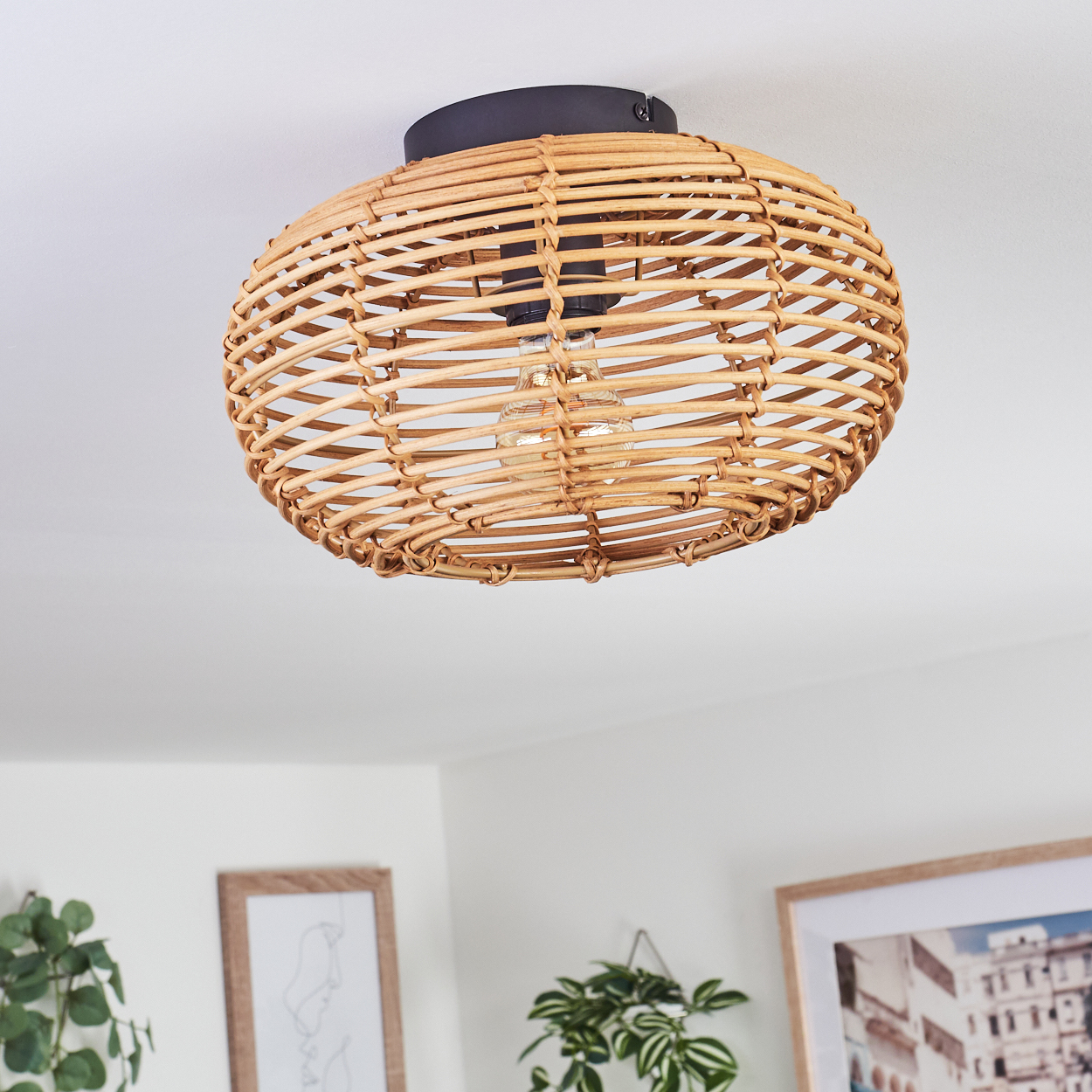 Eine gewebte Rattan-Leuchte hängt von der Decke. Darunter befinden sich gerahmte Kunstwerke und Pflanzen an einer weißen Wand.