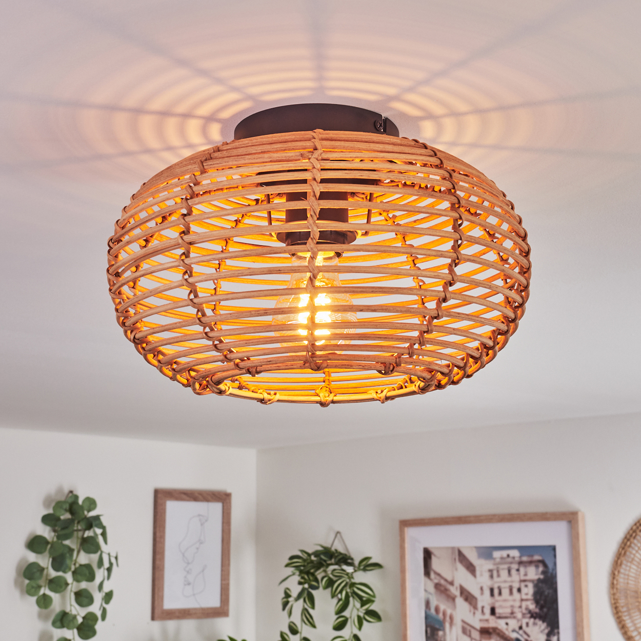 Eine runde Deckenlampe aus geflochtenem Rattan mit einer leuchtenden Glühbirne. Das Licht wirft einen Schatten auf die Decke. Gerahmte Kunst und Pflanzen im Hintergrund.