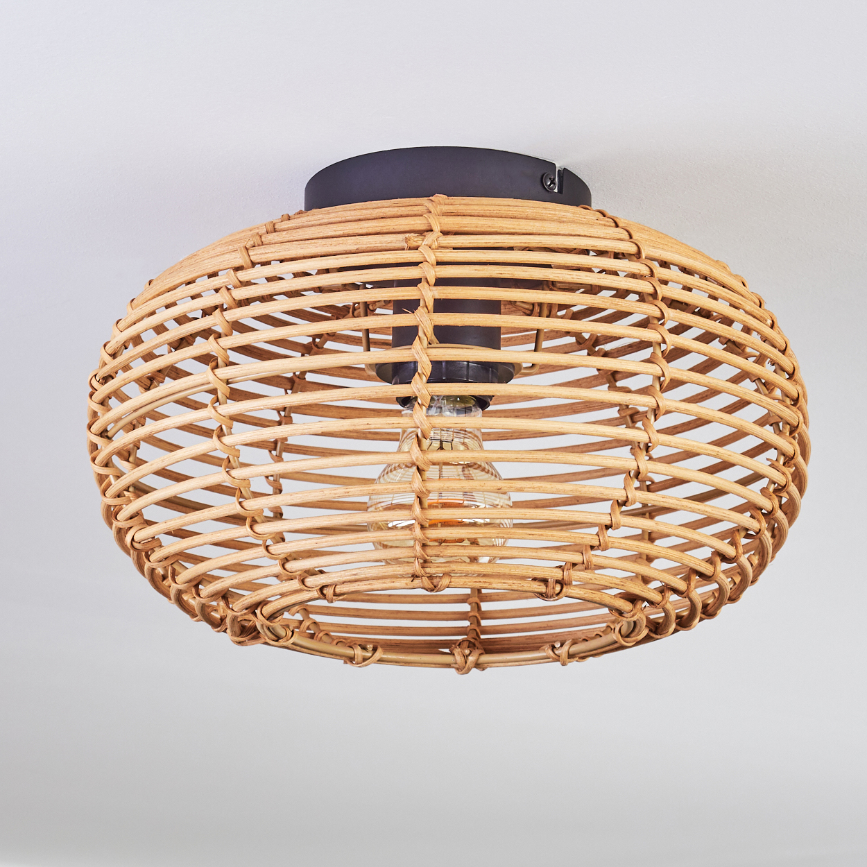 Eine gewebte Rattan-Leuchte hängt von einer weißen Decke. Sie hat einen schwarzen Sockel und eine sichtbare Glühbirne.