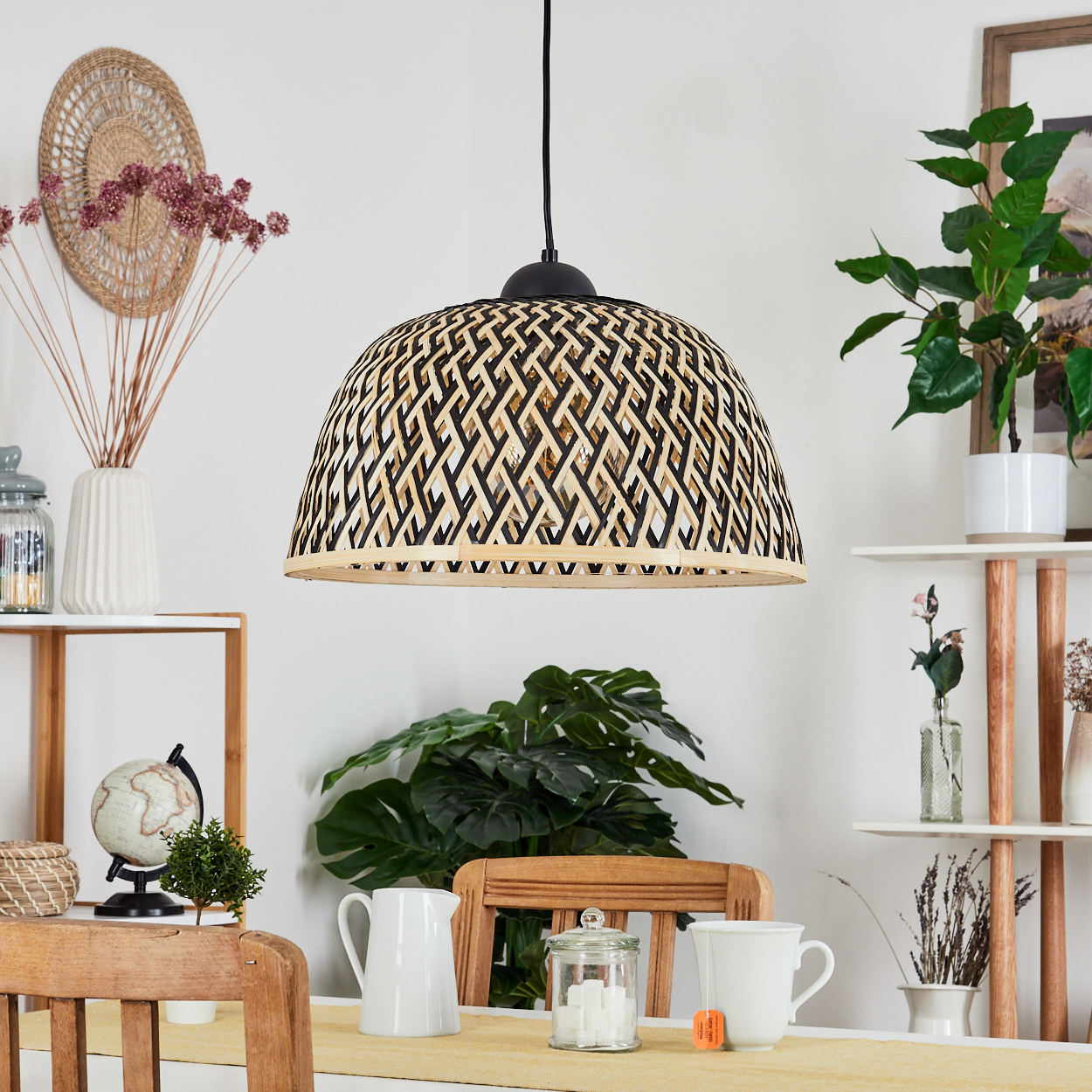 Ein gewebter Lampenschirm hängt über einem Holztisch, der mit einer weißen Kanne, einer Teetasse und einem Glas aufgedeckt ist. Pflanzen und Regale sind im Hintergrund.