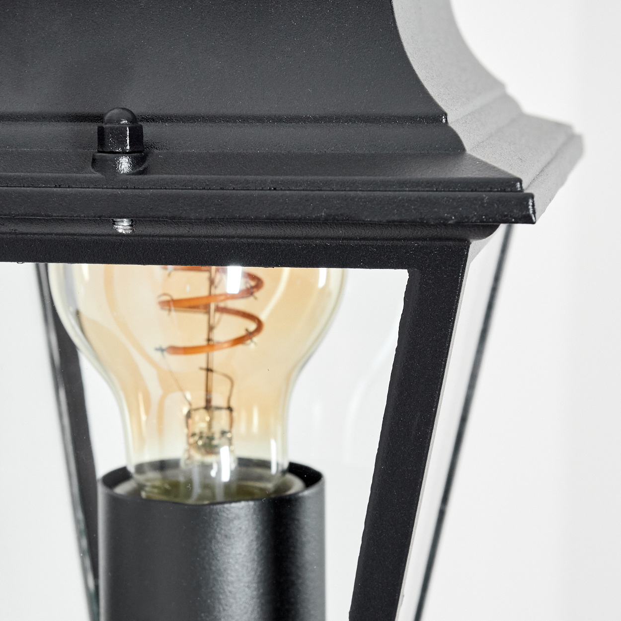 Nahaufnahme einer schwarzen Außenlampe. Eine leuchtende Glühbirne mit Wendel ist durch Glasscheiben sichtbar. Der Hintergrund ist eine weiße Wand.