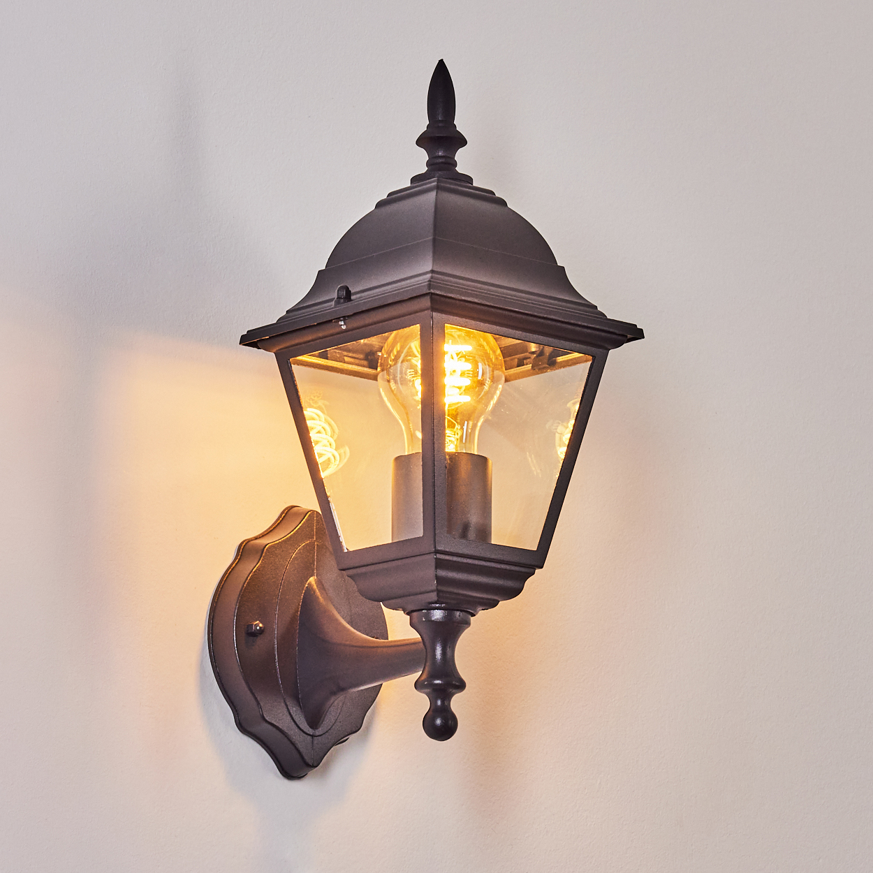 Eine schwarze, verzierte Außenlampe ist an einer hellbeige Wand angebracht. Die Lampe hat eine leuchtende Glühbirne im Inneren.
