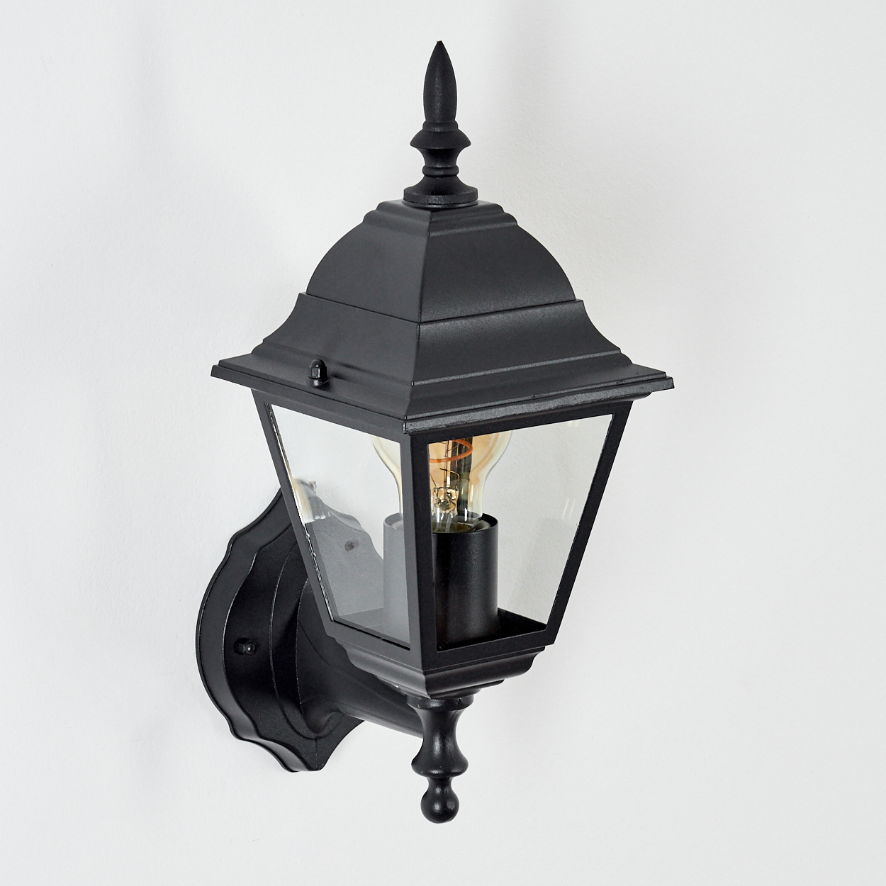 Eine schwarze Außenwandlampe mit Glasgehäuse und Glühbirne. Sie ist an einer weißen Wand montiert.