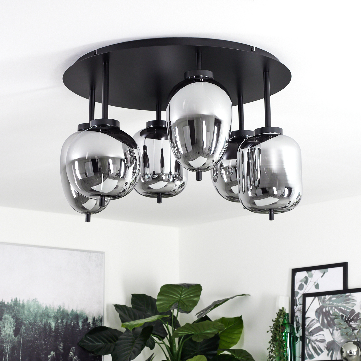 Eine moderne Deckenleuchte mit sechs reflektierenden, tropfenförmigen Lampen hängt von einer schwarzen, kreisförmigen Basis. Darunter sind Pflanzen und gerahmte Kunst zu sehen.