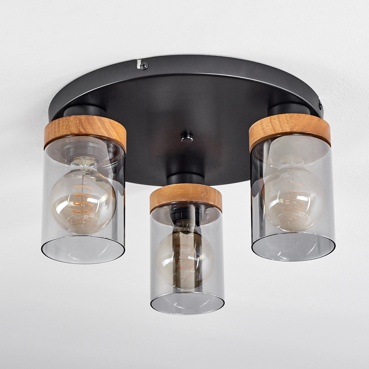 Deckenlampe mit drei Lichtern. Jedes Licht hat einen Glasschirm, einen Holzrahmen und eine sichtbare Glühbirne. Die Basis ist schwarz, die Decke weiß.