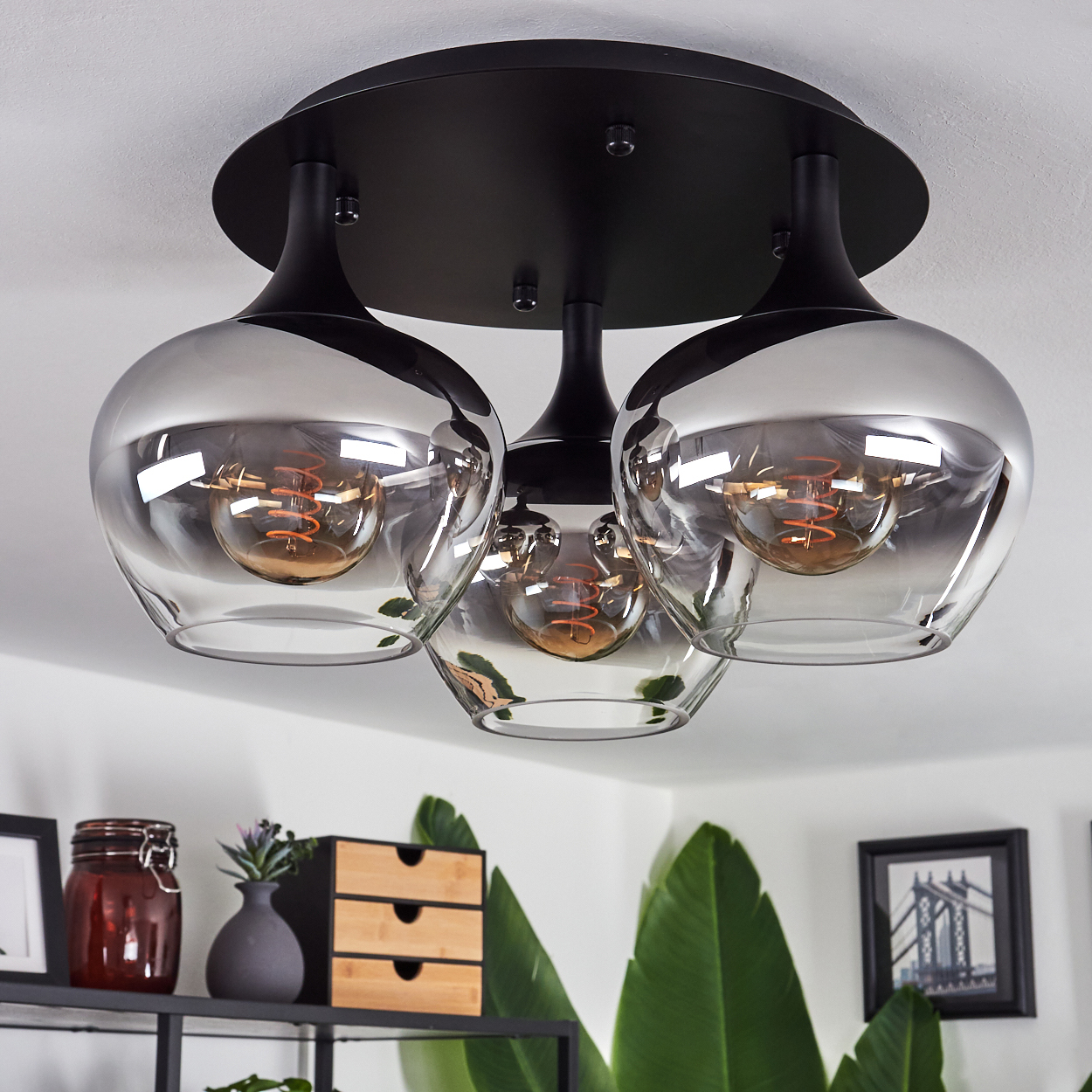 Eine moderne Deckenlampe mit drei Glasschirmen. Ein Regal darunter hat Deko, darunter eine kleine Holzschublade, eine Vase und gerahmte Kunst.