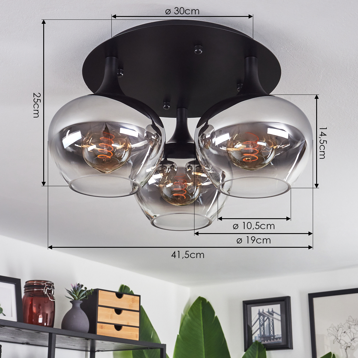 Eine schwarze Deckenlampe mit drei Glasschirmen, die jeweils eine Glühbirne enthalten. Abmessungen sind angegeben. Regal mit Dekoration im Hintergrund.